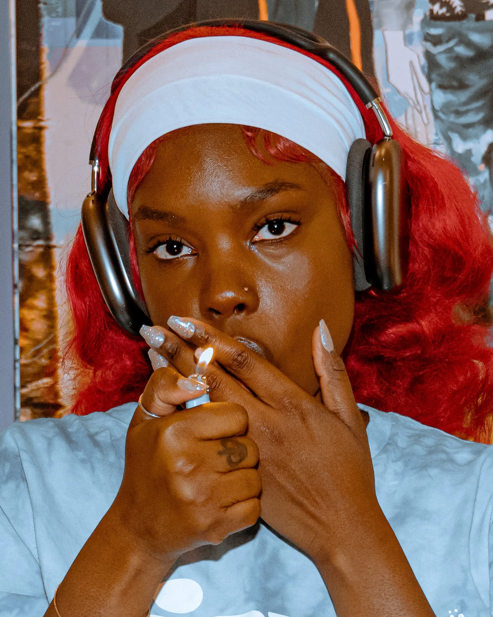 A woman with red hair and a nose ring wearing large headphones, holding a lit cigarette to her lips with her hands close to her face, and looking directly at the camera.