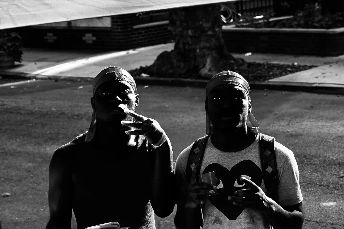 Two young men standing outdoors at night. One is wearing a sleeveless top and a durag, making a hand gesture near his face. The other is wearing a T-shirt with a heart design, a backpack, and a durag, holding a cup and making a gesture with his hand. The background features a tree, sidewalk, and a building.