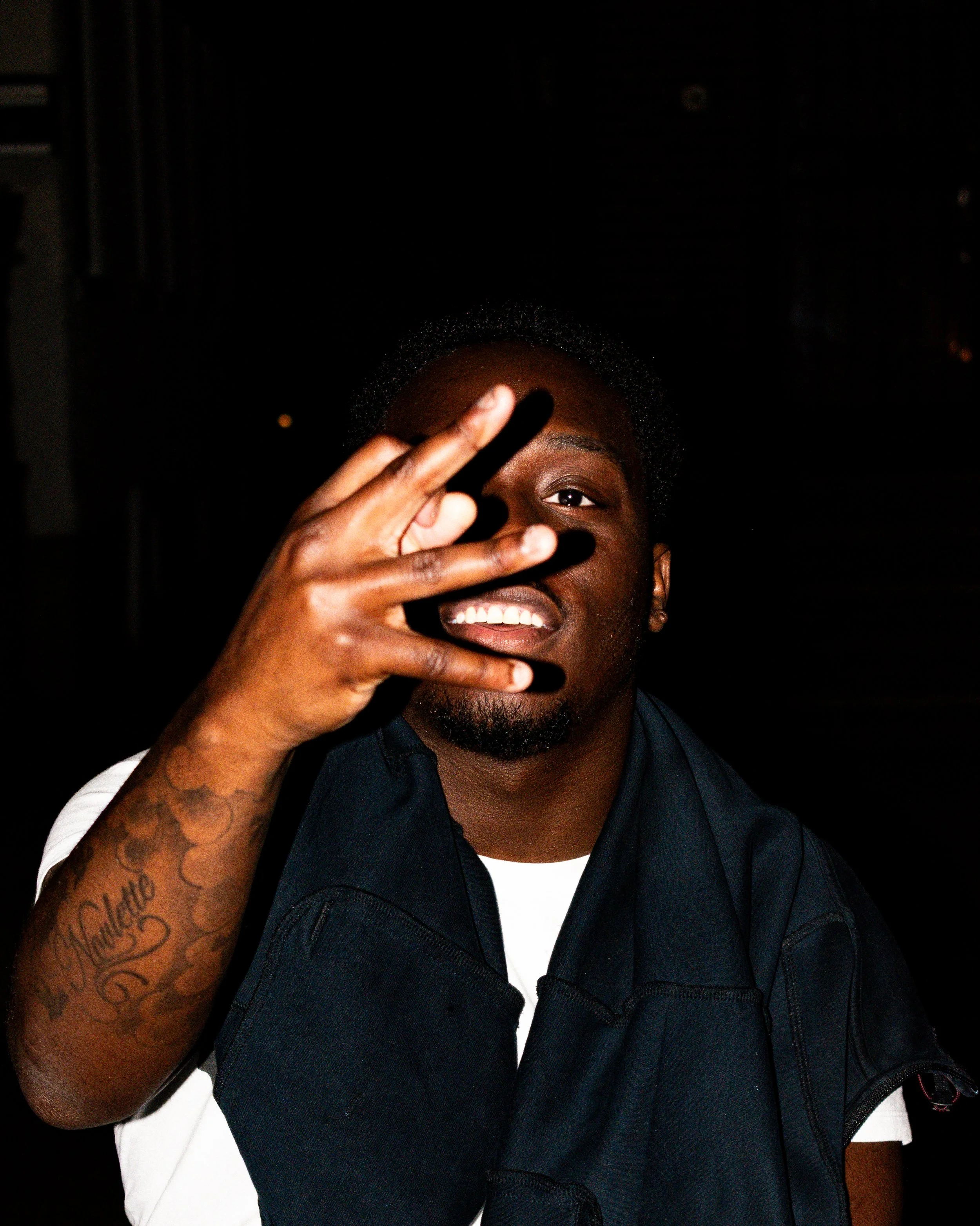 A young man with dark skin and tattoos on his arm, smiling and making a gesture with his hand in front of his face, against a dark background.