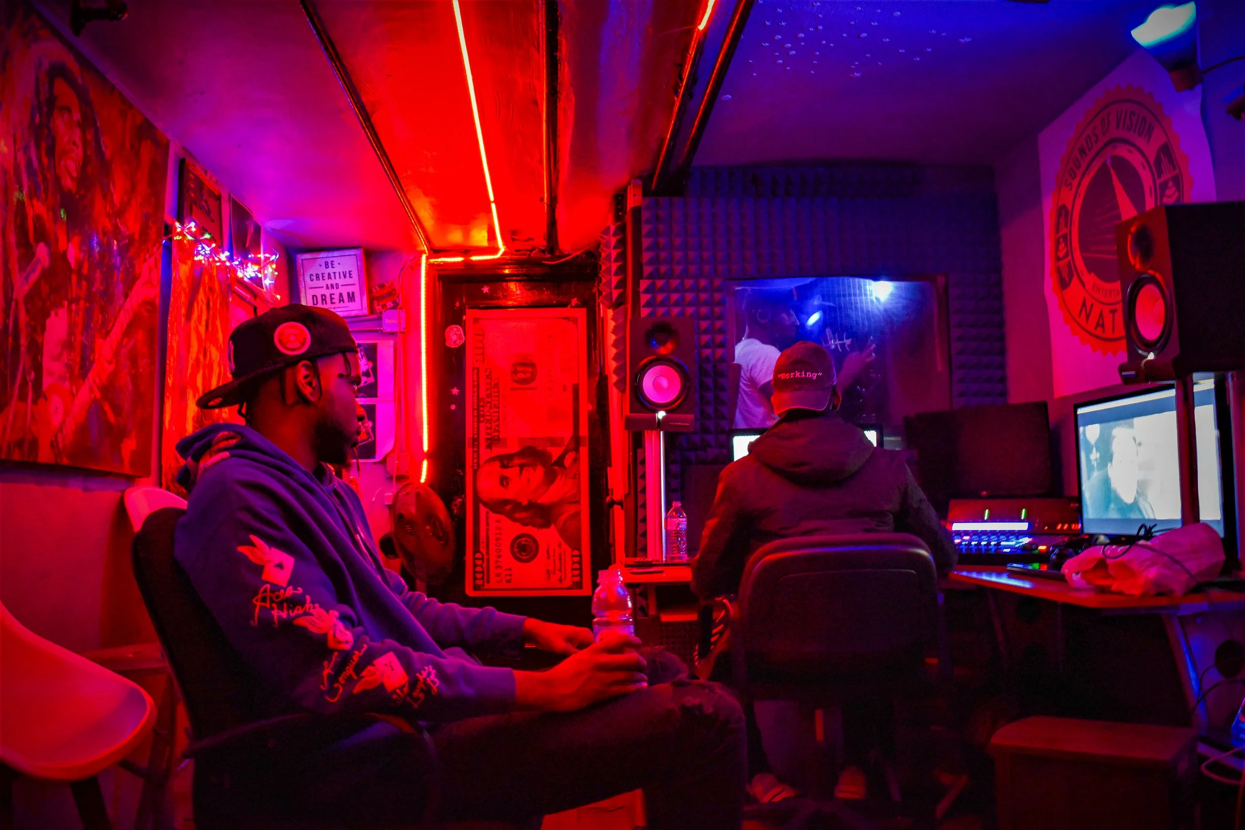Three people in a music studio or recording room with neon and colored lighting. One person is sitting holding a water bottle, two others are at computer workstations, with one seen through a glass window. The room is decorated with posters, art, and audio equipment.
