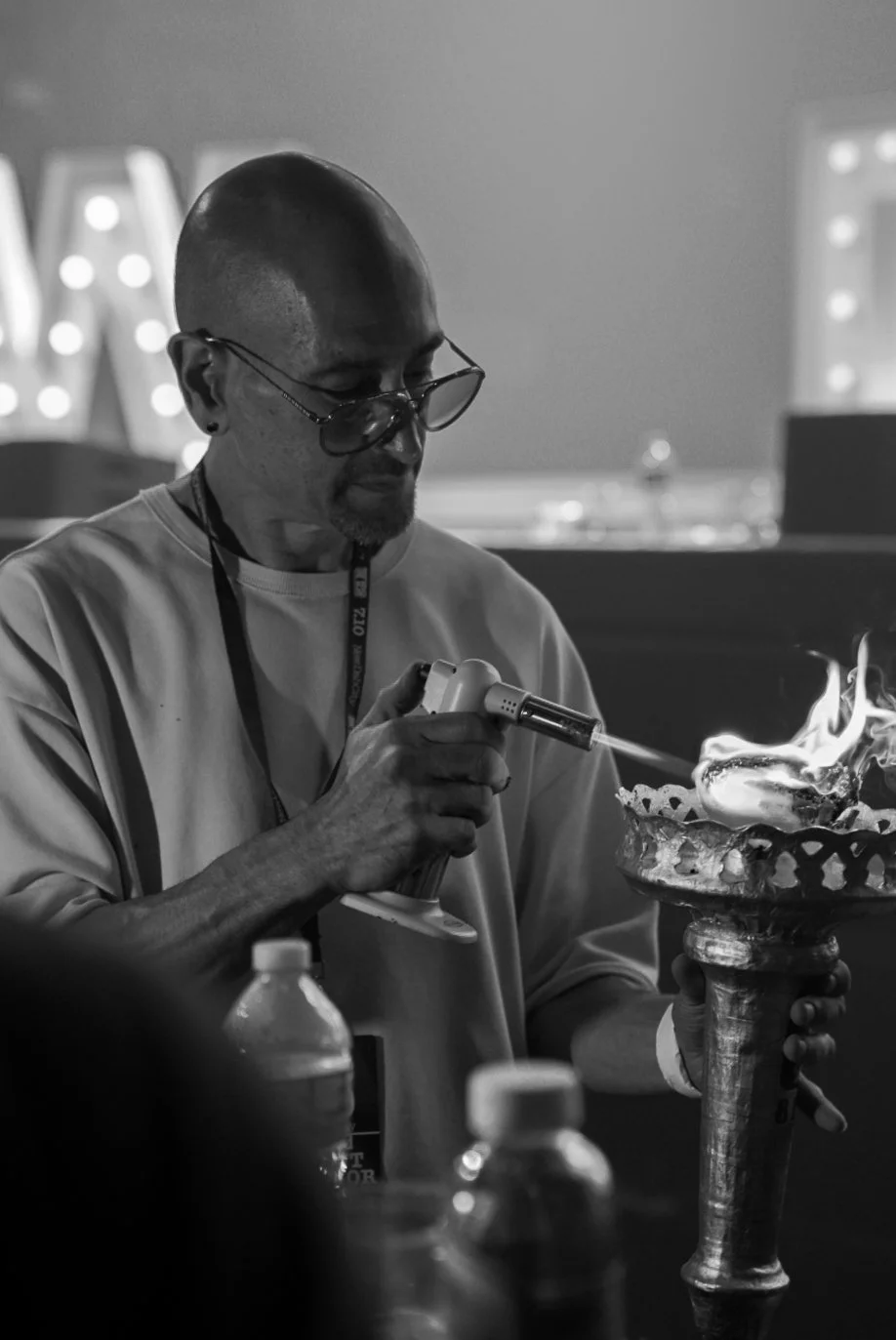 A man with glasses performing a flame demonstration, using an aerosol torch to create a fire display in a large decorative vessel.