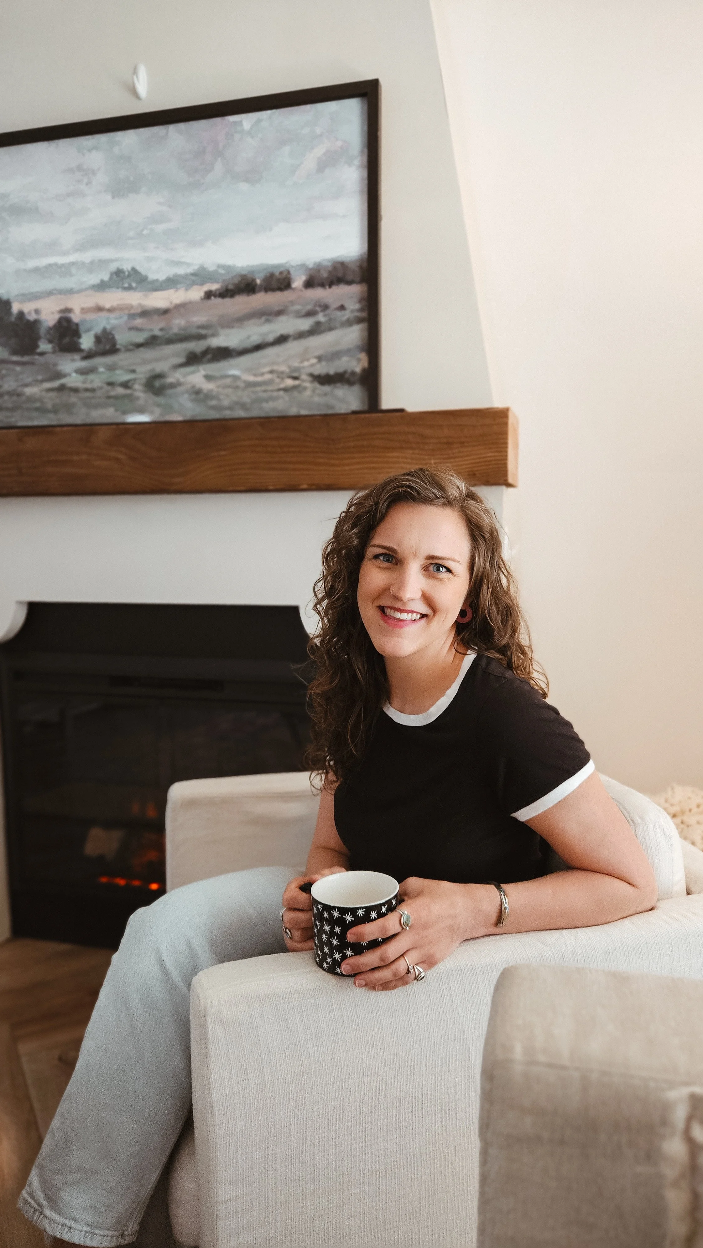 A woman with curly brown hair, wearing a black t-shirt with white trim, is sitting on a beige armchair by a fireplace, holding a black and white mug and smiling at the camera.