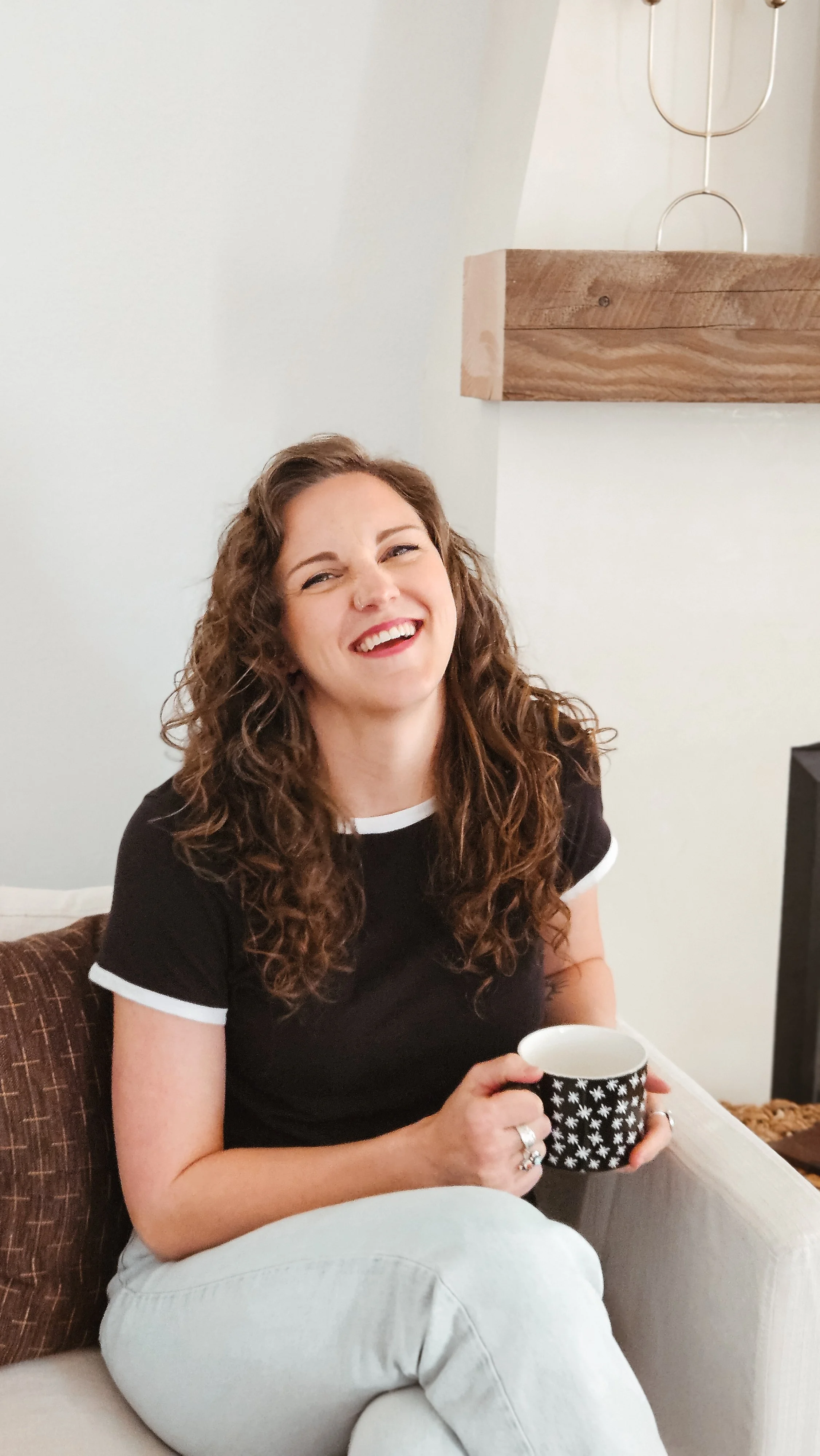 A woman with curly brown hair smiling and holding a black mug with white star pattern, sitting on a beige couch.