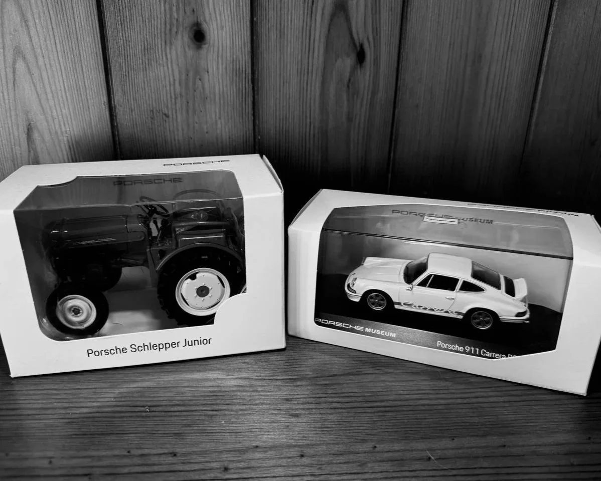 Porsche model cars displayed as collector pieces, reflecting modern classic automotive heritage, one is a Porsche Schleppar Junior tractor, and the other is a Porsche 911 Carrera RSR.