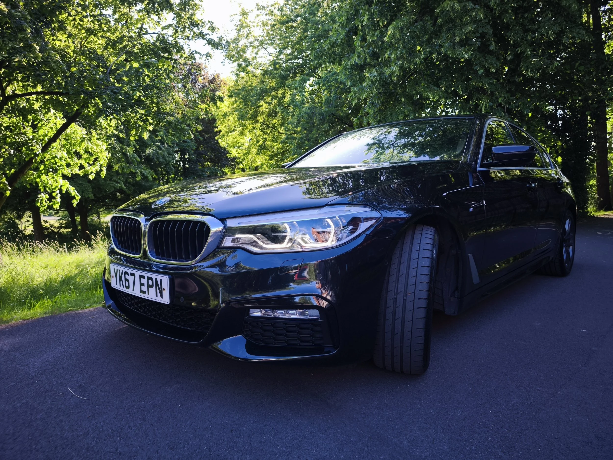 A shiny black BMW sedan parked on a paved surface in a lush wooded area with green trees and leaves overhead.