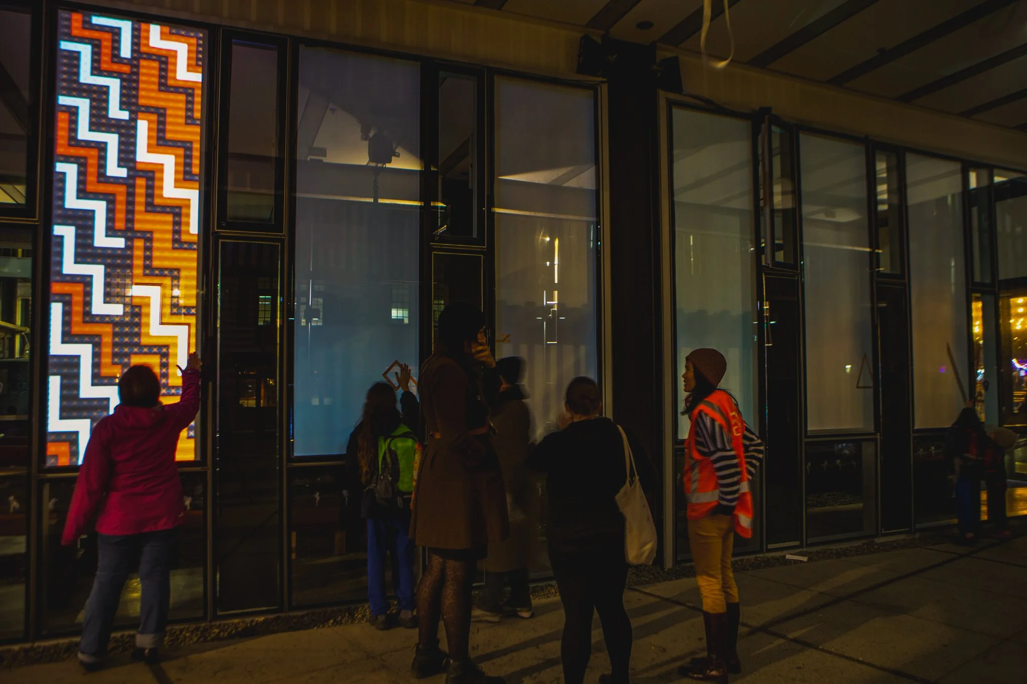 People gathered outside a building with large glass windows, some looking inside. One person is pointing at the window. The building has a colorful zigzag illuminated sign on the left side.