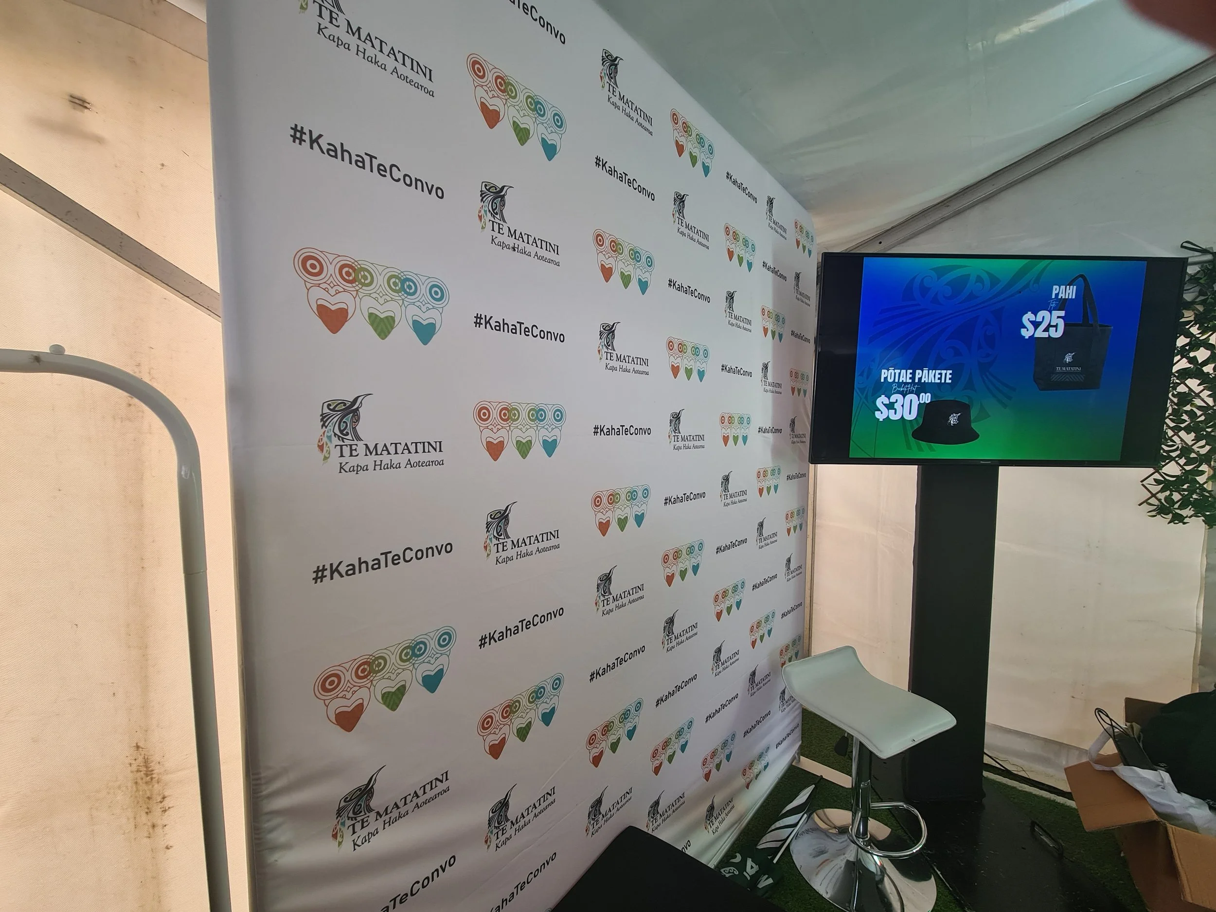 A promotional display with a repeating logo that features a stylized face with heart-shaped eyes and colorful design elements, and inclusive text, alongside a digital screen showing product prices of $25 and $30 for bags and hats.