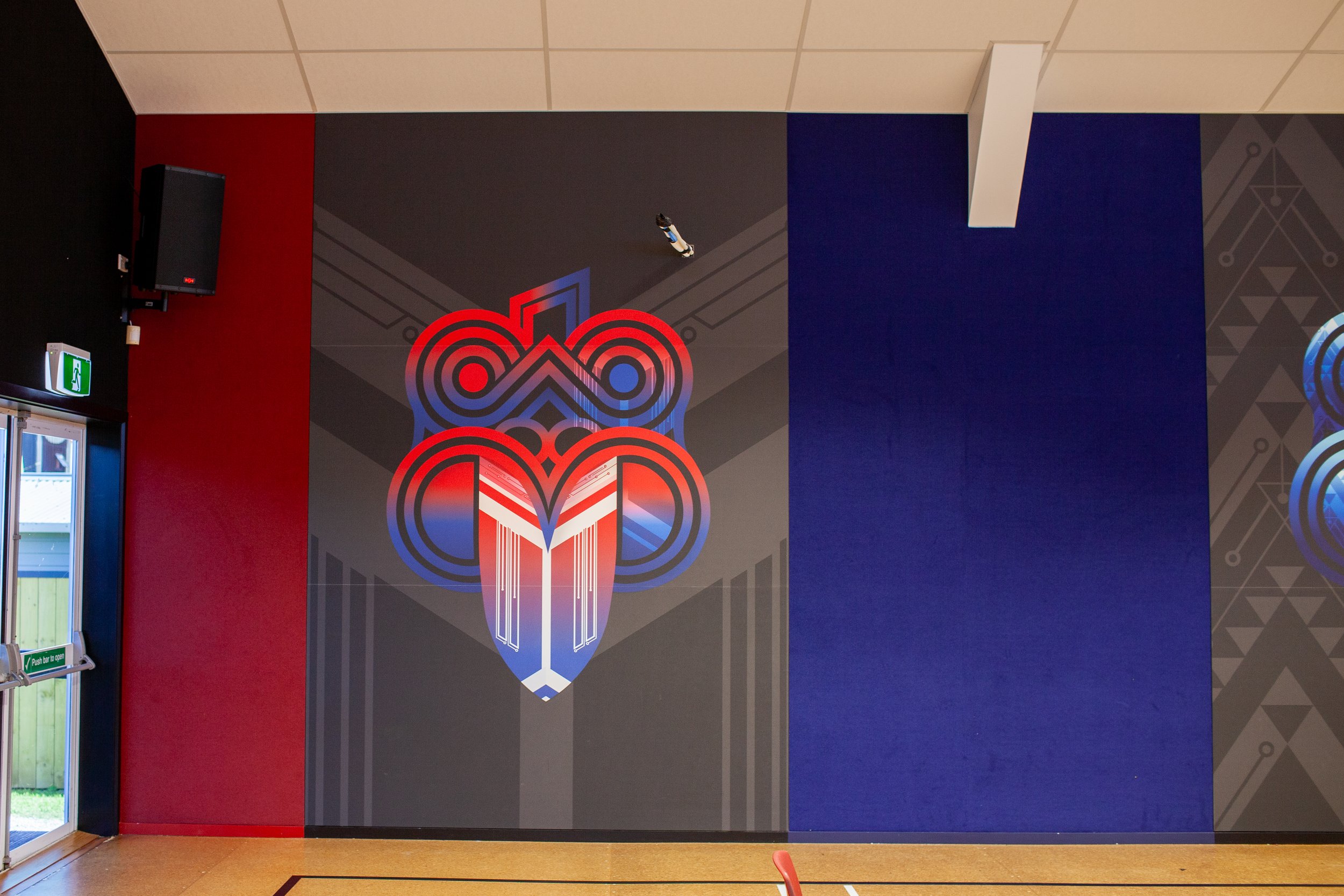 Interior wall with a colorful geometric mural design, featuring a large symmetrical tribal-inspired pattern in red, blue, and black on a gray background, with a section of dark blue and patterned gray adjacent. A black speaker is mounted in the upper left corner and a green emergency exit sign with a white running figure is above the glass door to the outside.