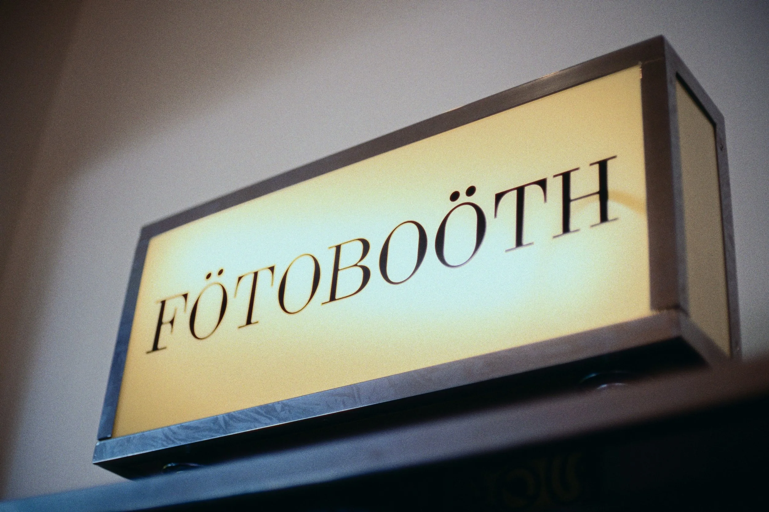 Illuminated sign with the word 'FÖTOBOÖTH' in black letters on a yellow background, likely indicating a photo booth.