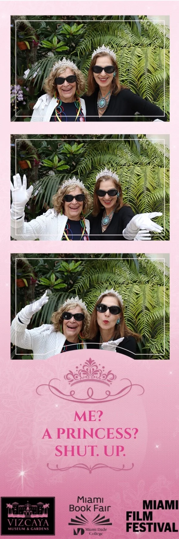 Two women wearing tiaras and sunglasses posing in a photo booth with a backdrop of tropical plants. One woman is waving and the other is making a puckered face. They are dressed in black and white clothing with colorful jewelry, and are wearing white