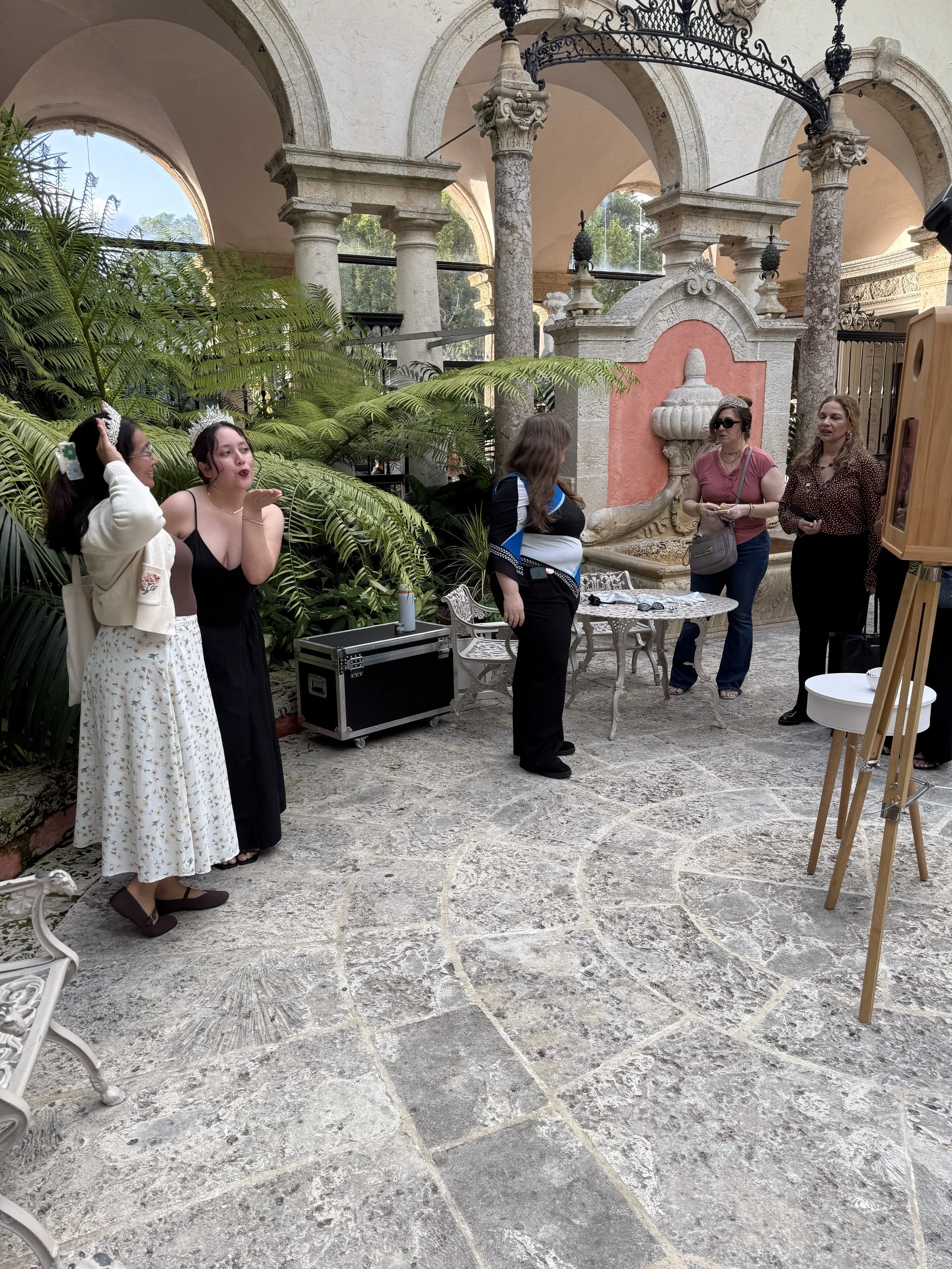 People gathered in an ornate courtyard with arches, columns, and a fountain, some with tiaras and casual attire, engaging in conversation or posing for photos.