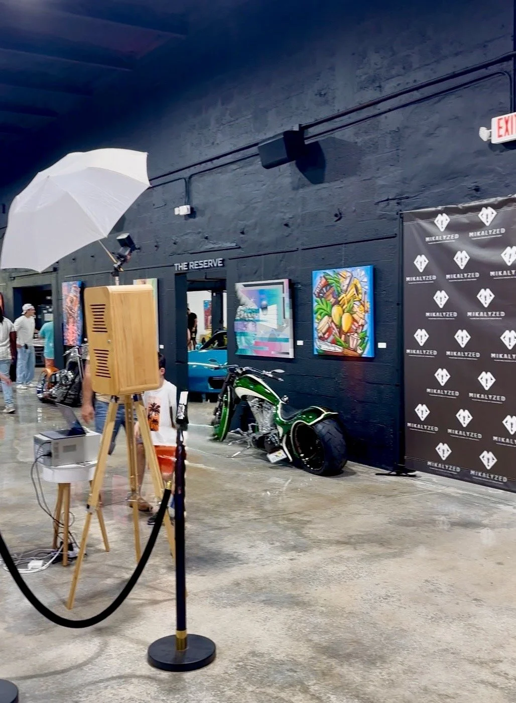 Art exhibition area with paintings on a dark wall, a motorcycle, and a car in the background. There are people walking and a portable light setup with a white umbrella.