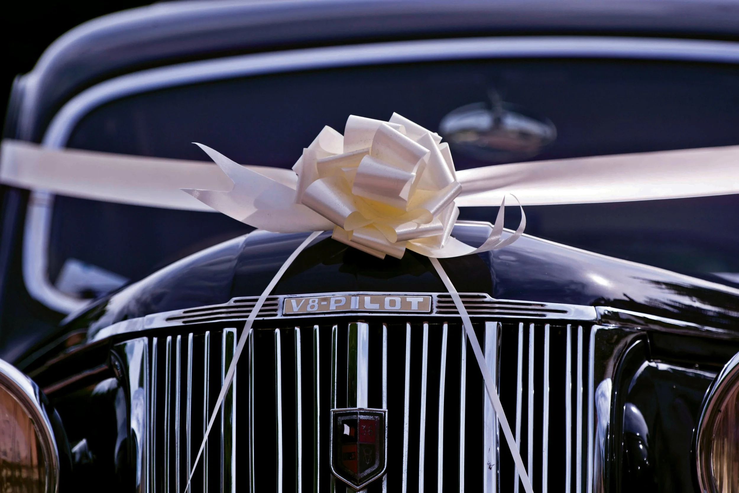 Close-up of a black vintage car with a large white ribbon and bow on the hood, indicating it is a wedding or special occasion vehicle.