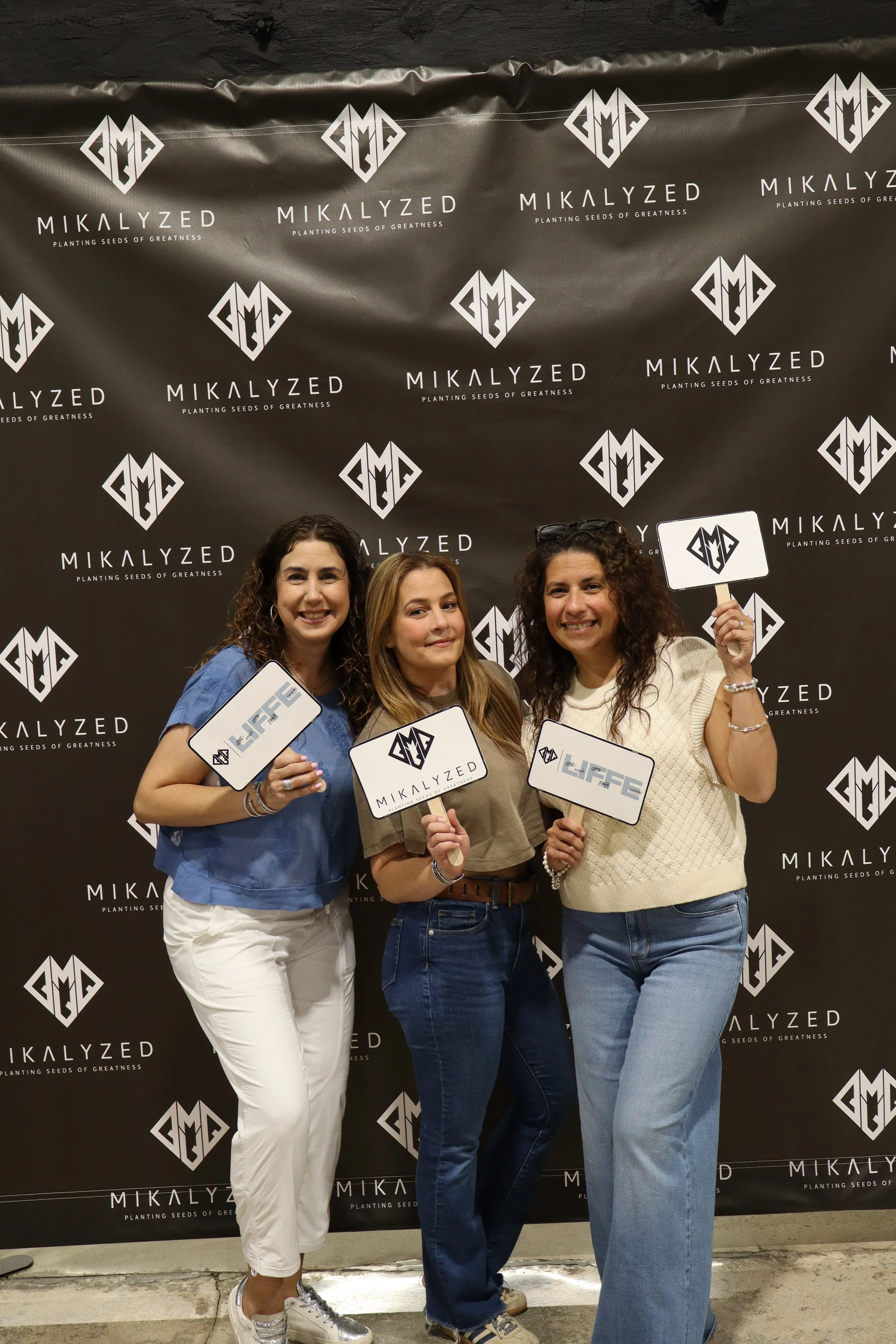 Three women standing in front of a black backdrop with white logos and text, smiling and holding signs that say 'Mikalzed' and 'Life'.