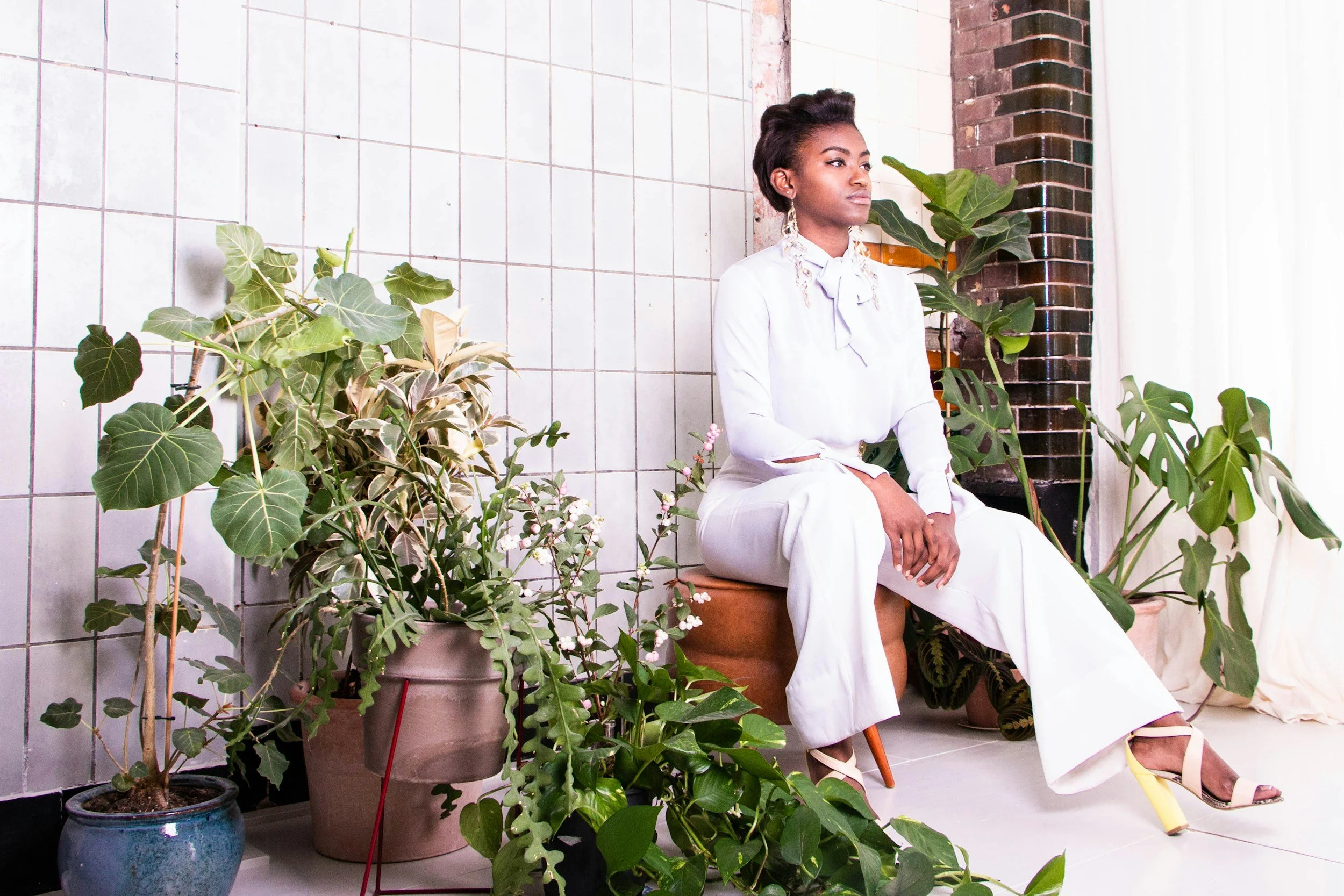 A woman sitting on a stool among lush green potted plants in a room with tiled walls and brick accent wall, gazing thoughtfully to the side.