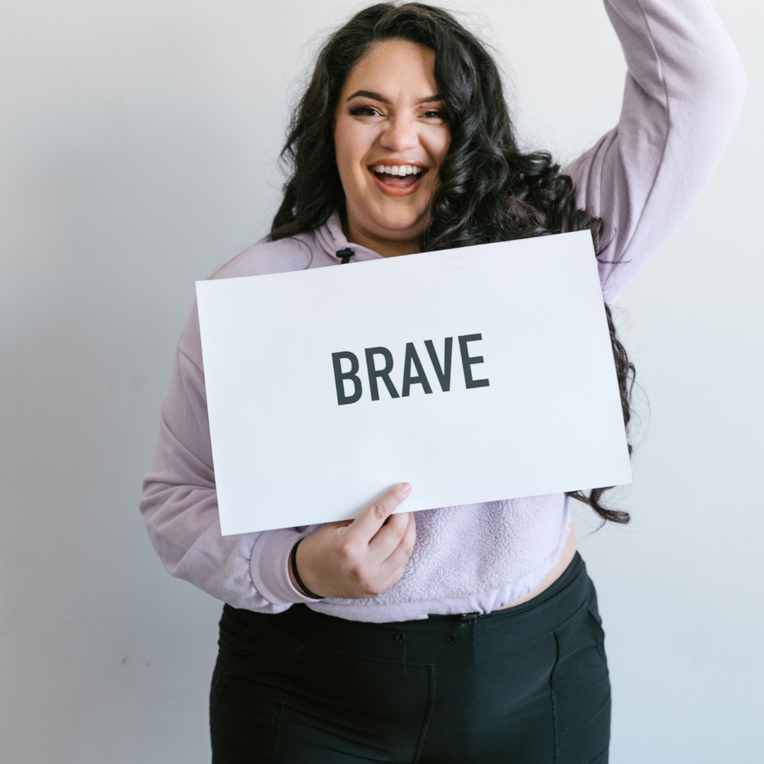 A Woman smiling with one hand in the air holding a sign that says brave story work-coaching-robrenna