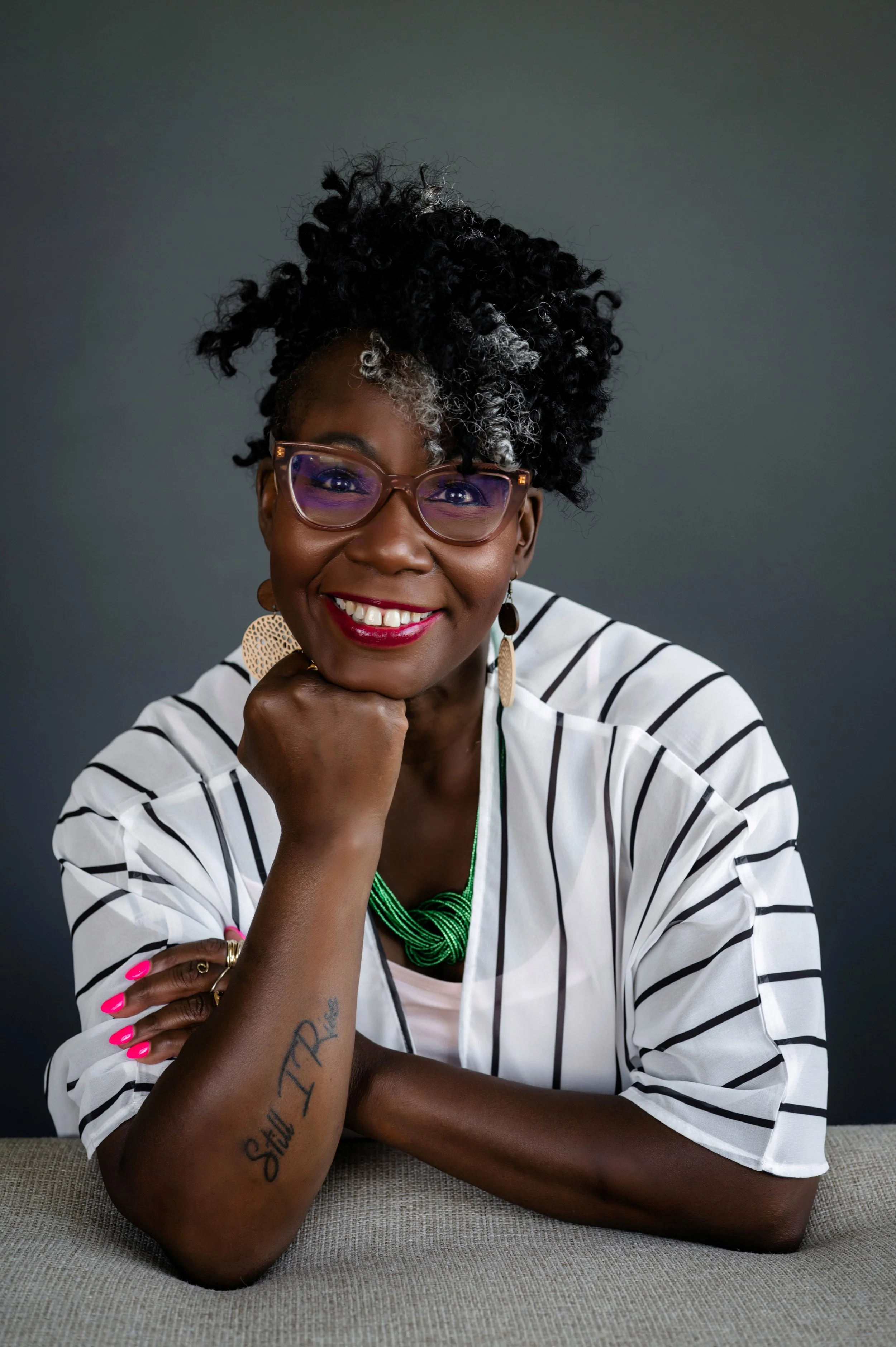 Smiling African American woman with curly hair short hair, wearing glasses, earrings, a white and black striped shirt, with colorful jewelry, sitting at a table with a gray background.