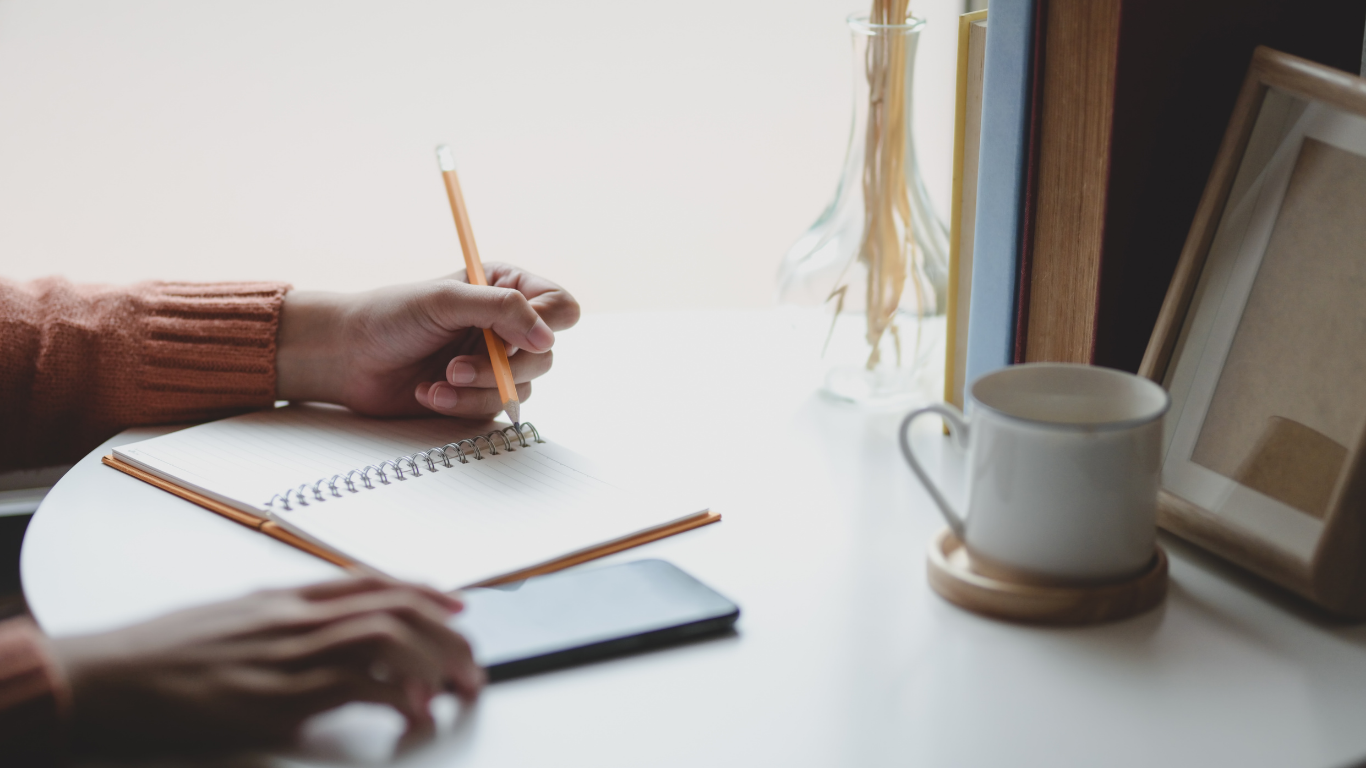 A person's hands writing in a notebook with a pencil in one hand and phone on table under other hand. A coffee mug on a coaster and a picture frame behind it.