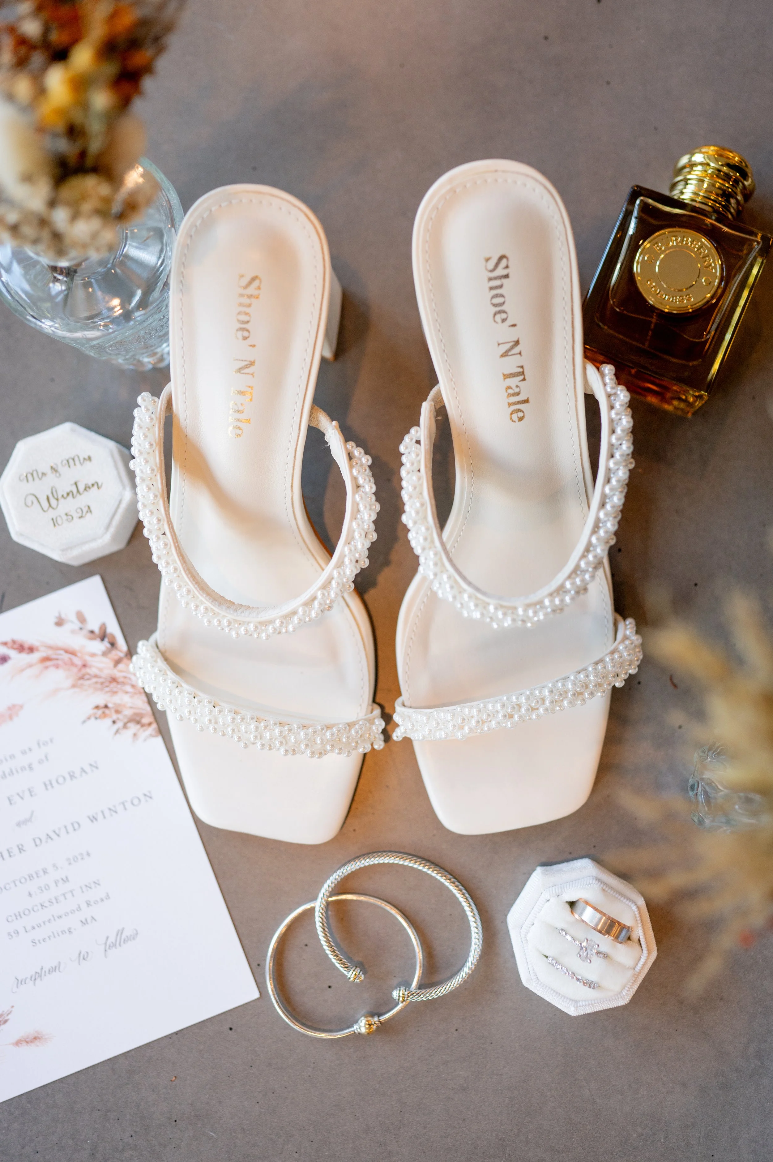 White embellished high-heeled shoes, wedding rings, perfume, jewelry, wedding invitation, and small decorative items for a wedding.