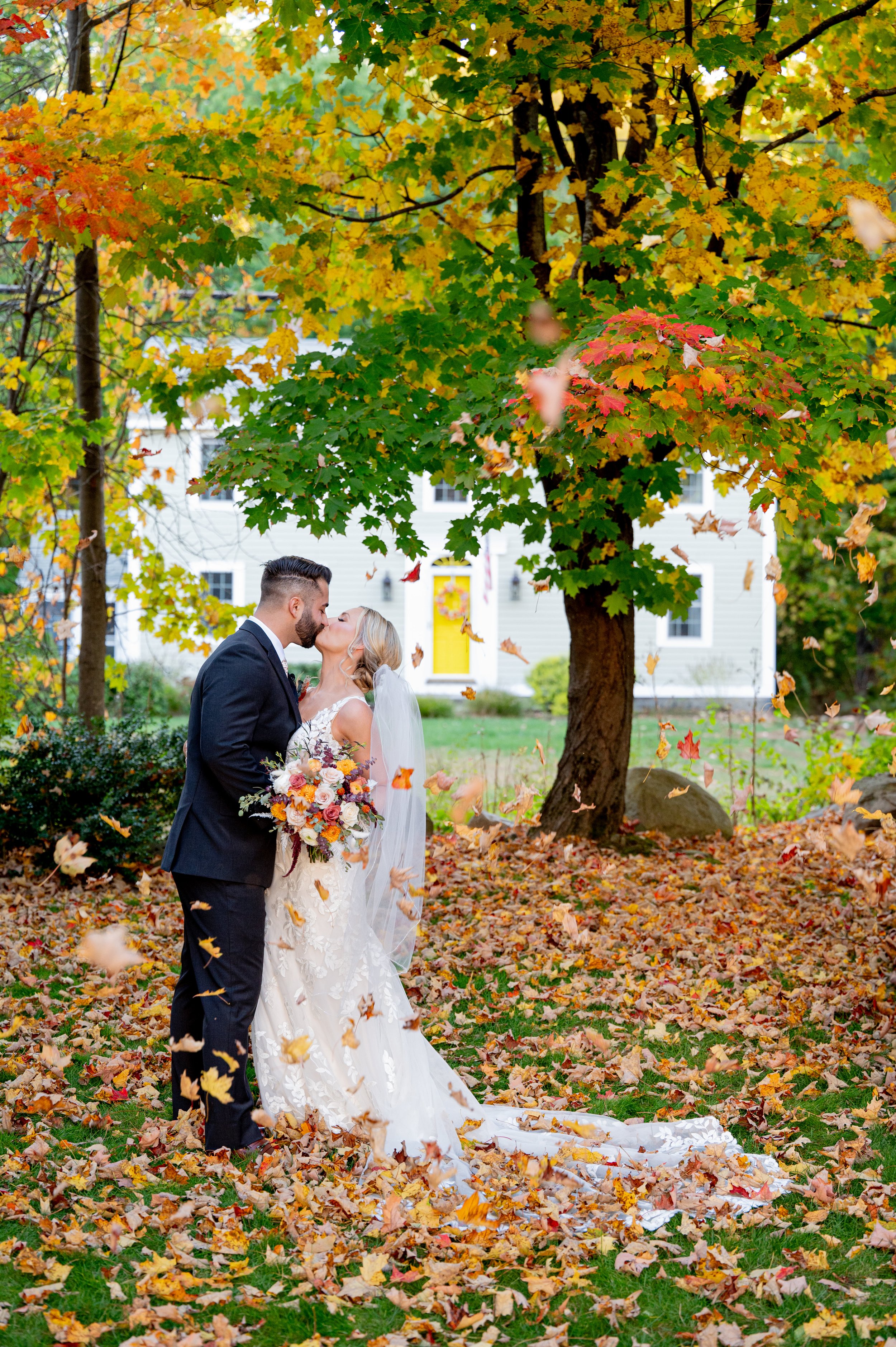 A bride and groom kissing under fall foliage in a park, surrounded by colorful fallen leaves, with a white house in the background.