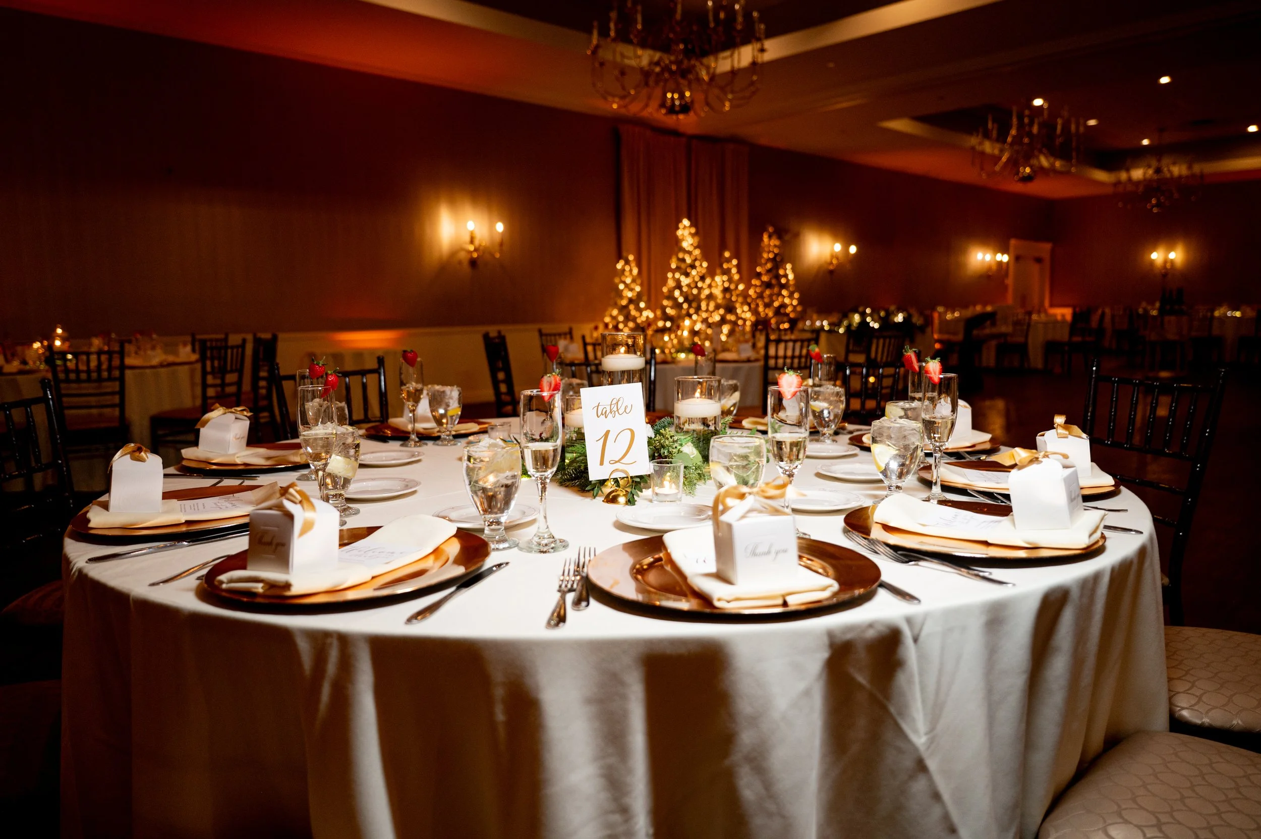 Elegant round table set for a formal event with gold-rimmed charger plates, white napkins, and small gift boxes with gold ribbons, surrounded by black chairs. In the center, a decorative arrangement of candles with a table number card reading 'table 