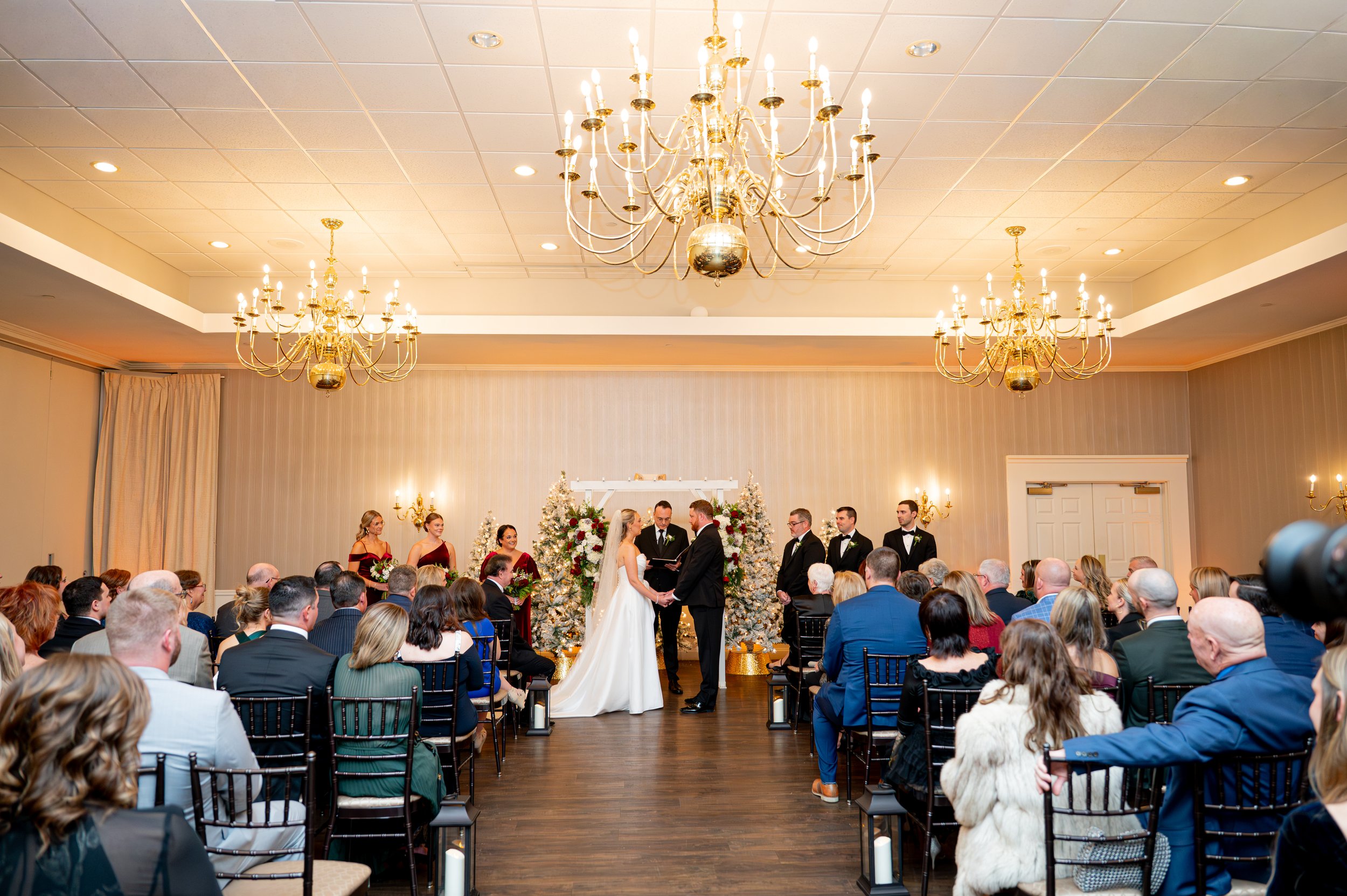Wedding ceremony in a decorated indoor venue with chandeliers, featuring a bride and groom exchanging vows in front of a seated audience, with a decorated altar and Christmas trees in the background.