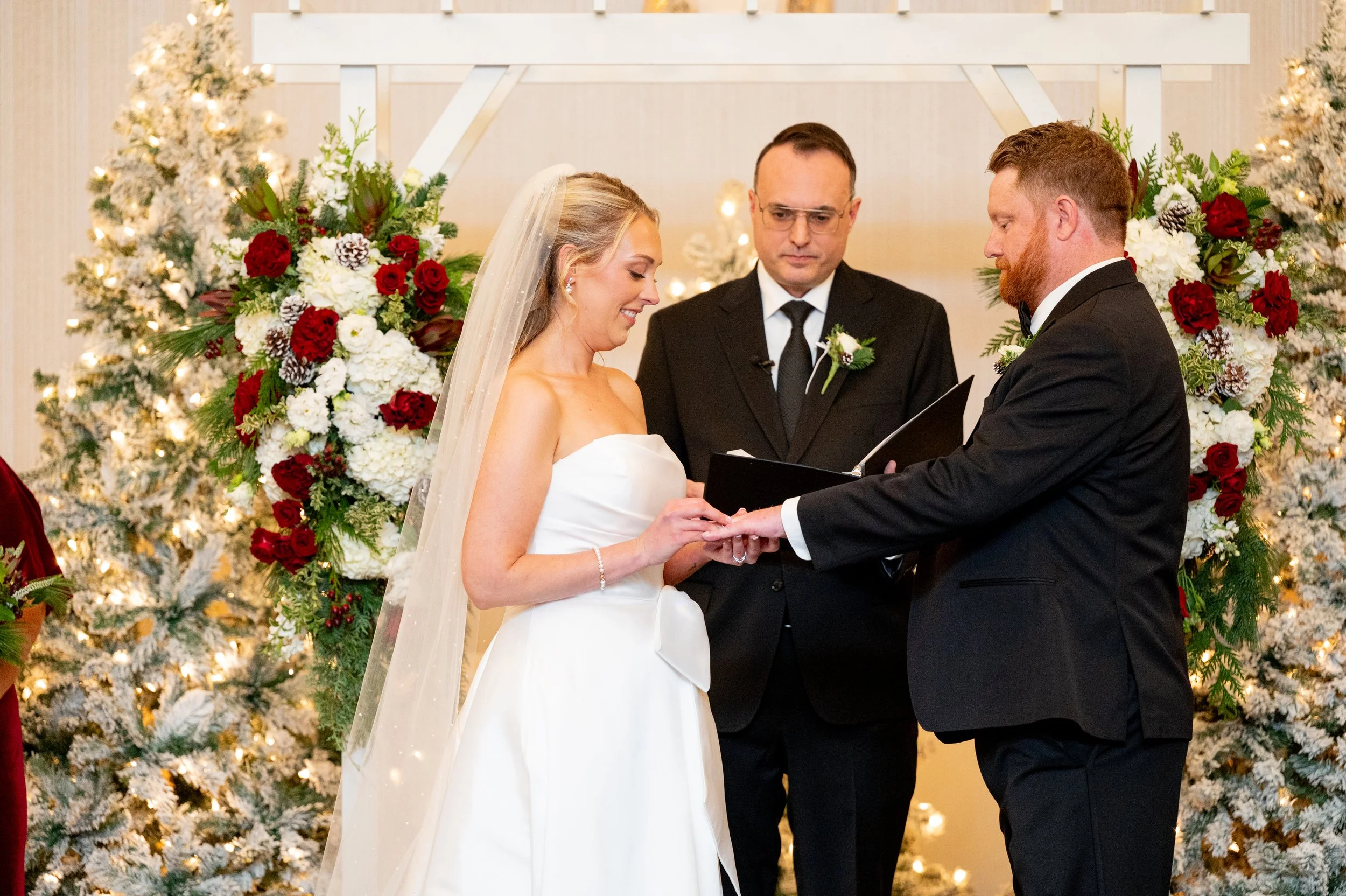 A bride and groom exchanging wedding rings during their wedding ceremony with an officiant standing behind them, in front of Christmas-themed decor with snowy white trees and red and white flowers.