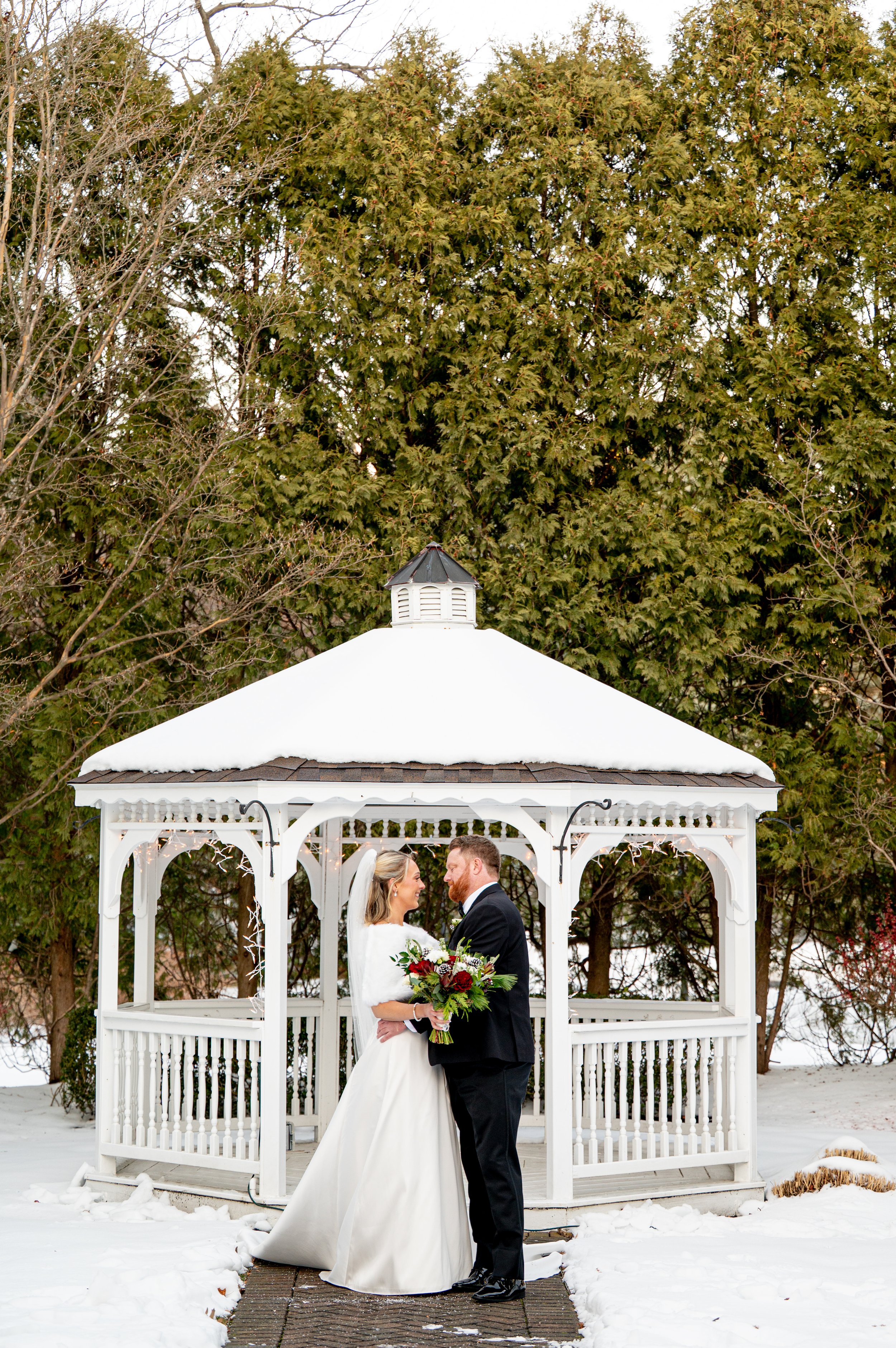 A bride and groom in wedding attire standing close together in front of a white gazebo with snow on top, holding a bouquet, and surrounded by snow and trees.