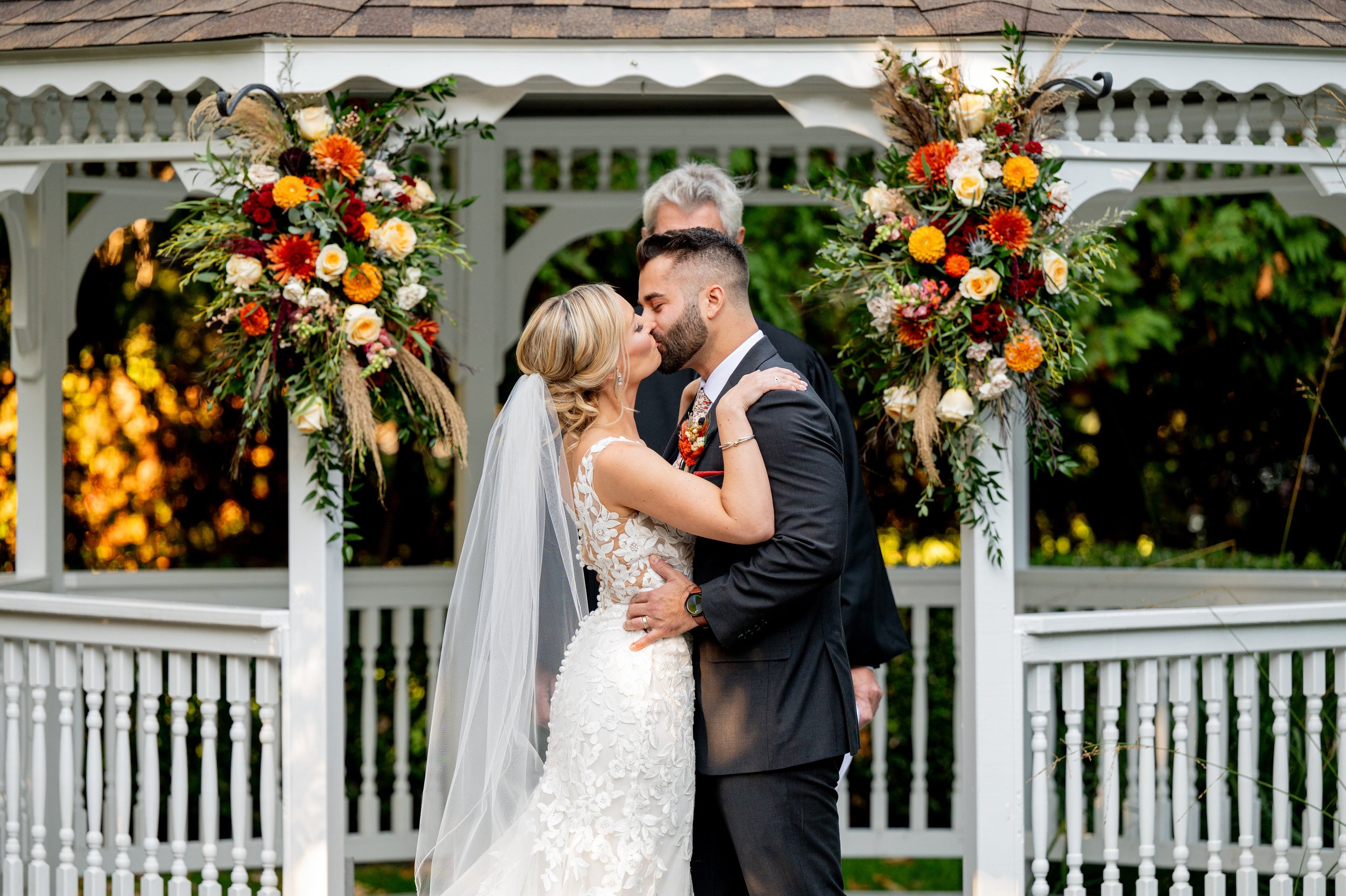A bride and groom sharing a kiss during their wedding ceremony under a decorated arbor with colorful flower arrangements, with a blurred background of trees and sunset lighting.