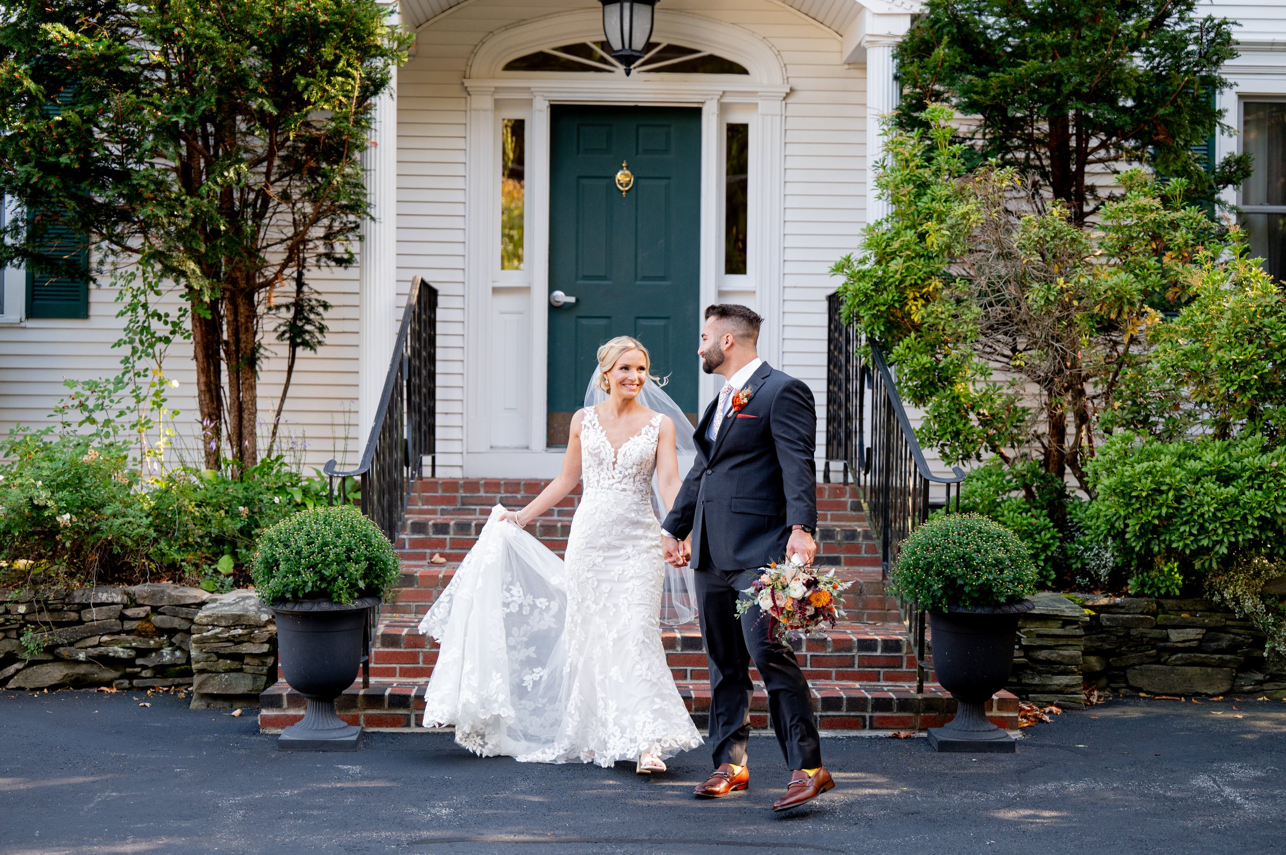 Bride and groom walking hand in hand down the driveway outside a house, smiling at each other. The bride is holding her wedding dress with one hand, and the groom is holding a bouquet of flowers in his other hand. The house has white siding, a teal front door, and brick steps with black railings, flanked by green bushes and potted plants.