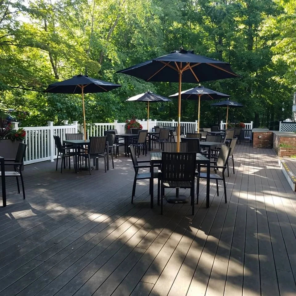 An outdoor patio area with several black tables and chairs, large black umbrellas, white railing, and lush green trees in the background.