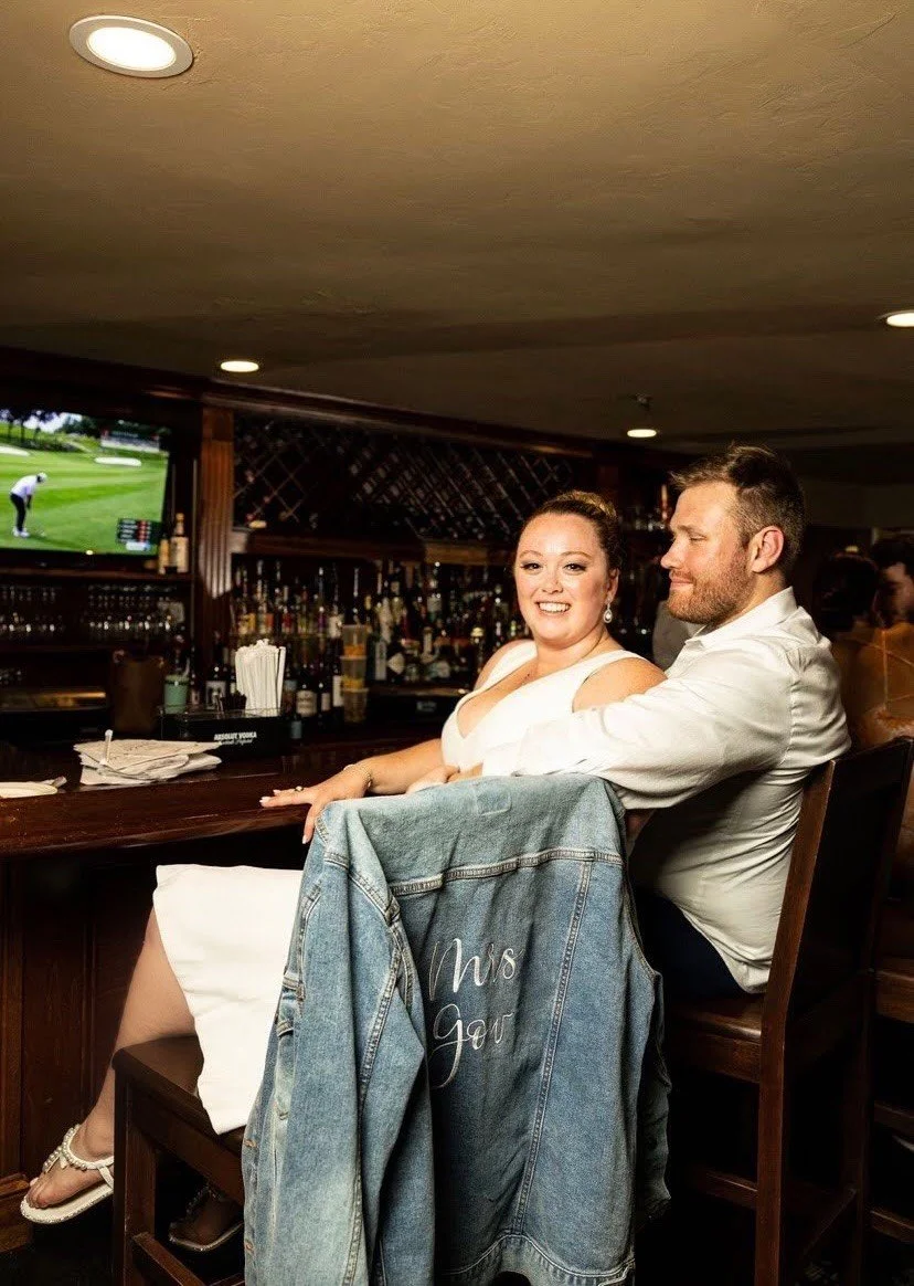 A smiling couple, a woman in a white dress and a man in a white shirt, sitting at a bar inside a restaurant or bar, with a large screen TV showing a golf game in the background. The woman has a denim jacket hanging on the back of her chair with the words "This is our" written on it.