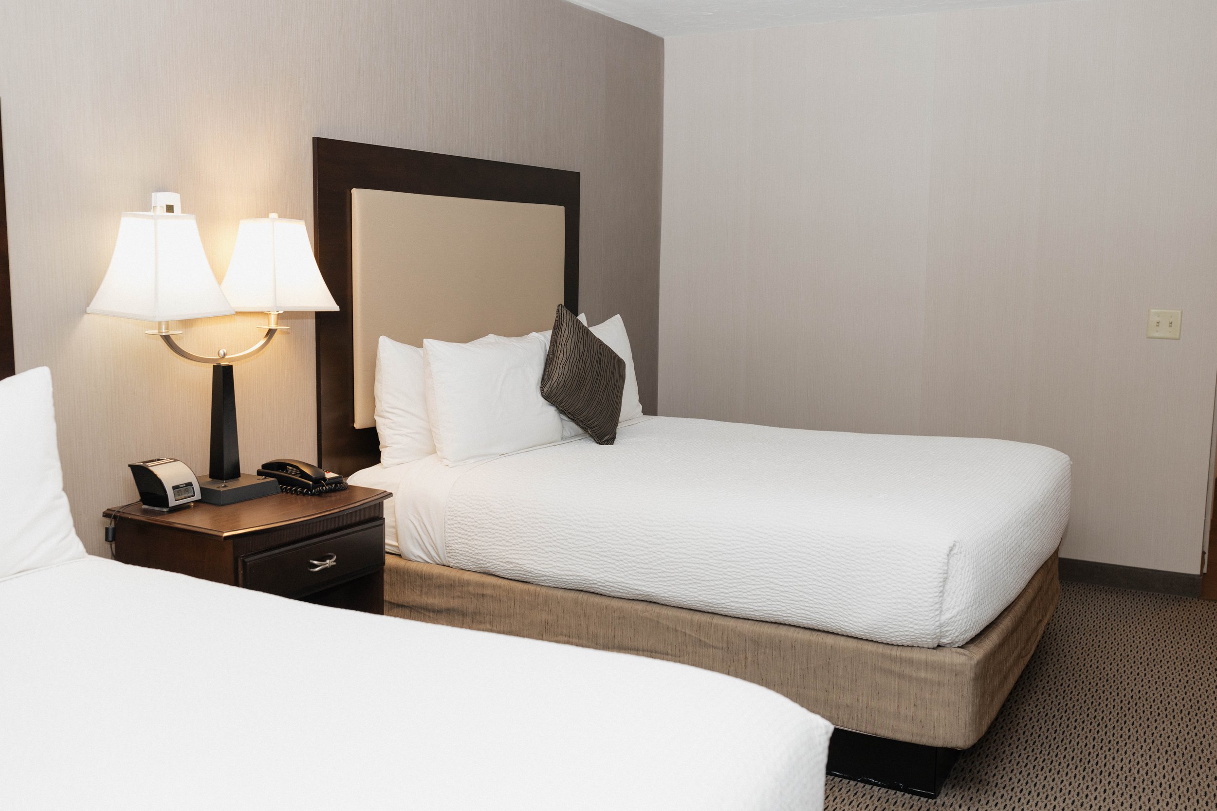 Hotel room with two beds, a nightstand with a lamp, telephone, and alarm clock between the beds, beige walls, and carpeted floor.