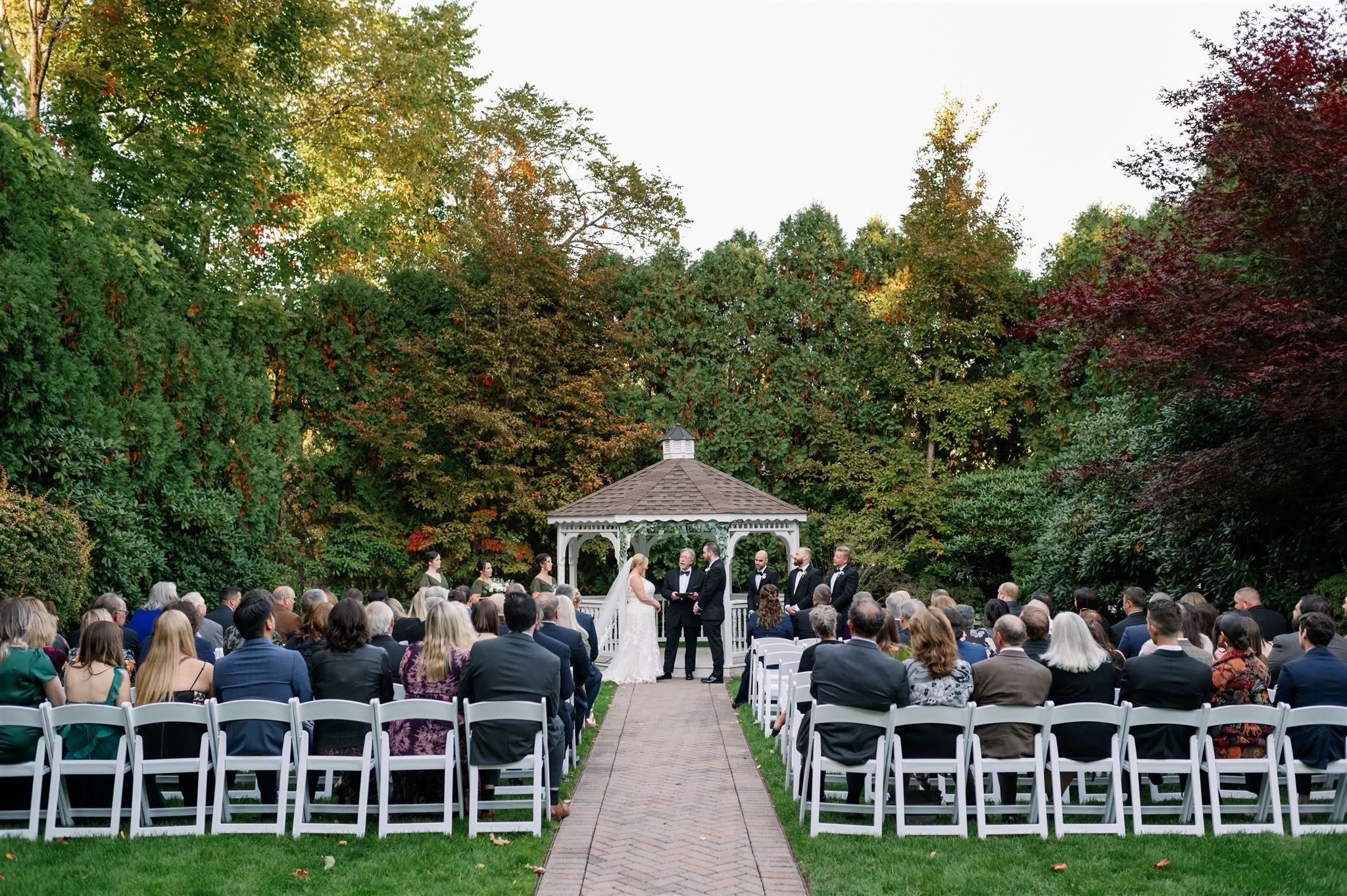 Outdoor wedding ceremony with guests seated on white chairs facing a gazebo, where a bride and groom stand with officiant and officiant is reading. The scene is surrounded by lush green trees with hints of autumn colors.