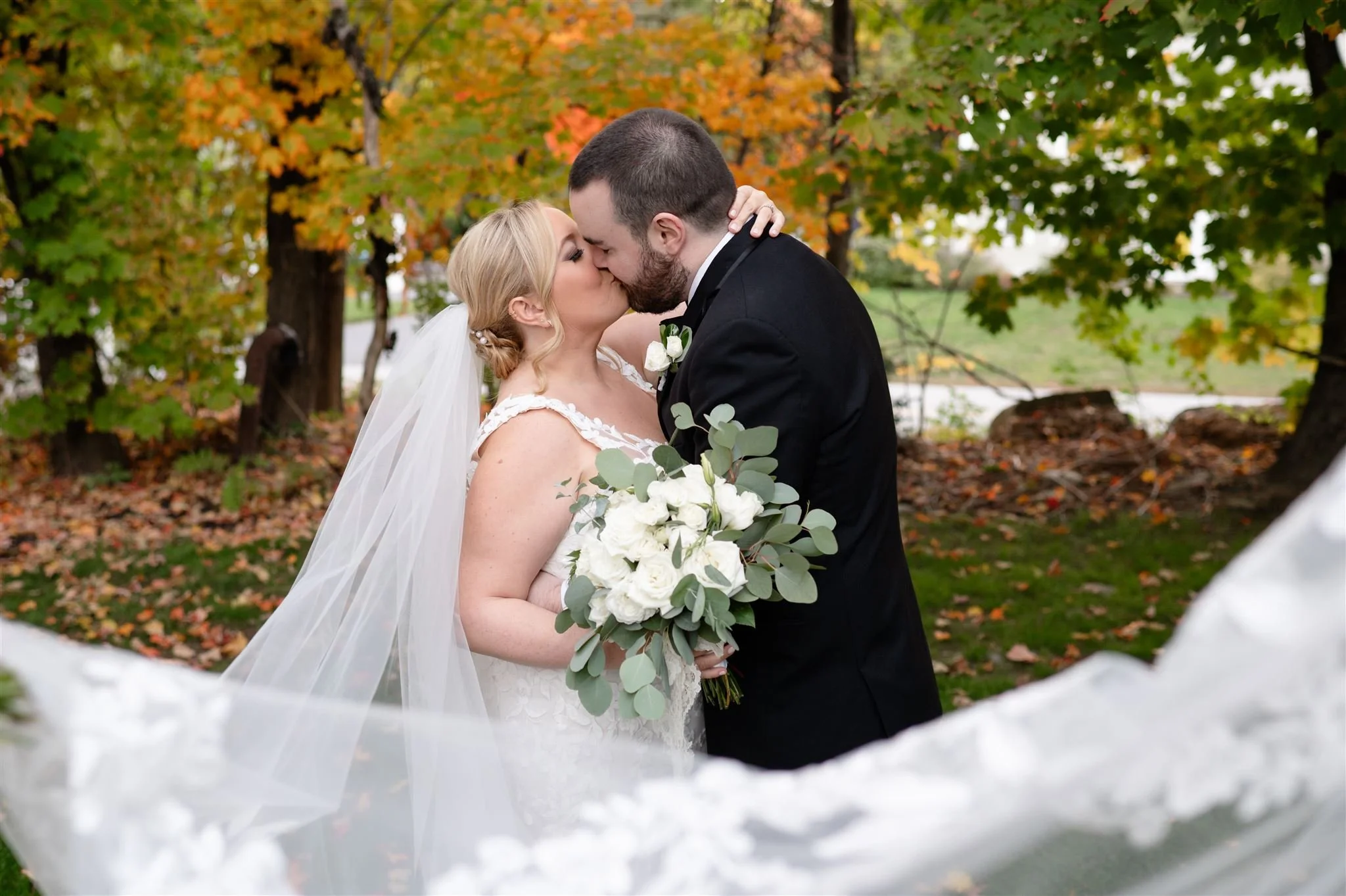 A bride and groom kiss outdoors in front of fall foliage; she holds a bouquet of white flowers and greenery.