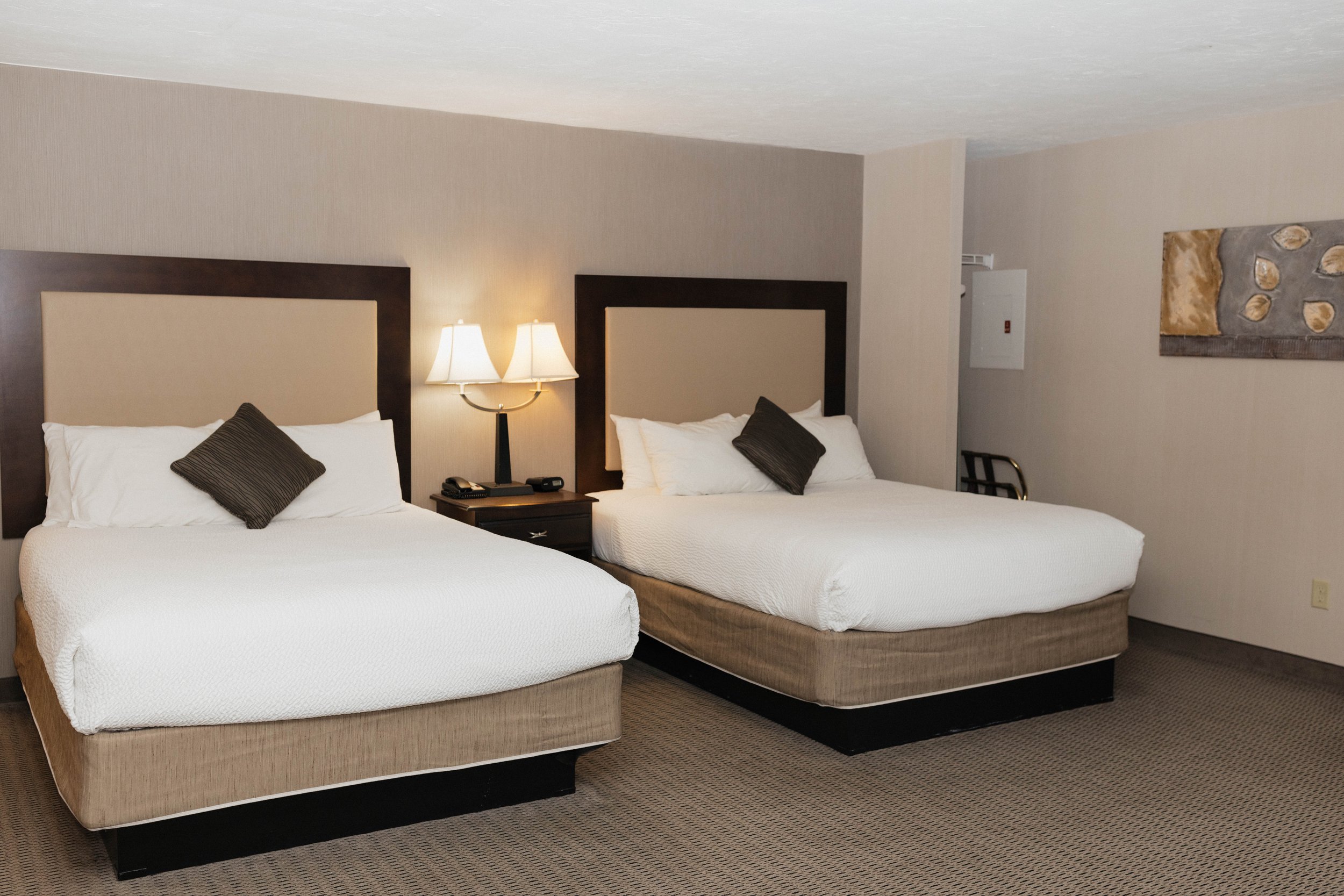 Two queen beds with beige headboards, white bedding, and pillows, a nightstand with a lamp and hotel phone, and a piece of abstract art on the wall.