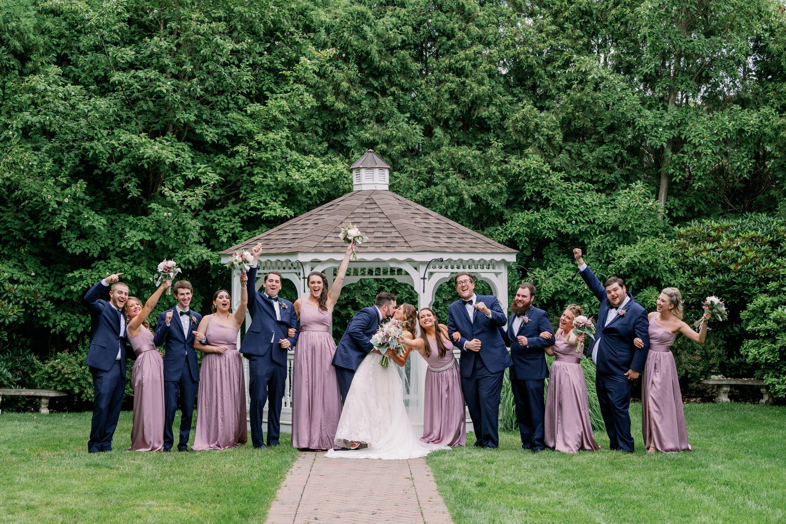 A joyful wedding party posing outdoors in front of a gazebo, with the bride and groom surrounded by bridesmaids and groomsmen, all smiling and celebrating, holding bouquets and raising their arms.