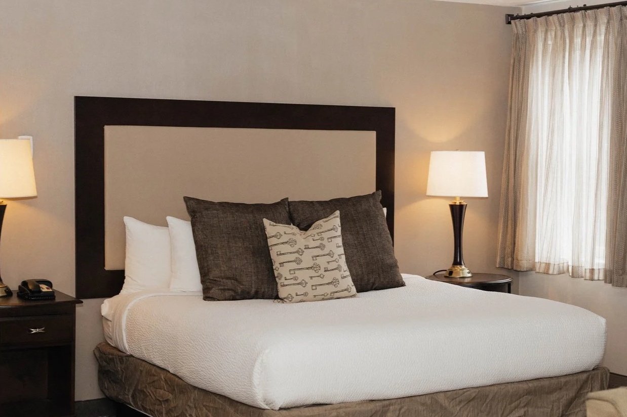 A neatly made hotel bed with white bedding and multiple pillows, two dark brown nightstands with lamps, and a window with beige curtains.