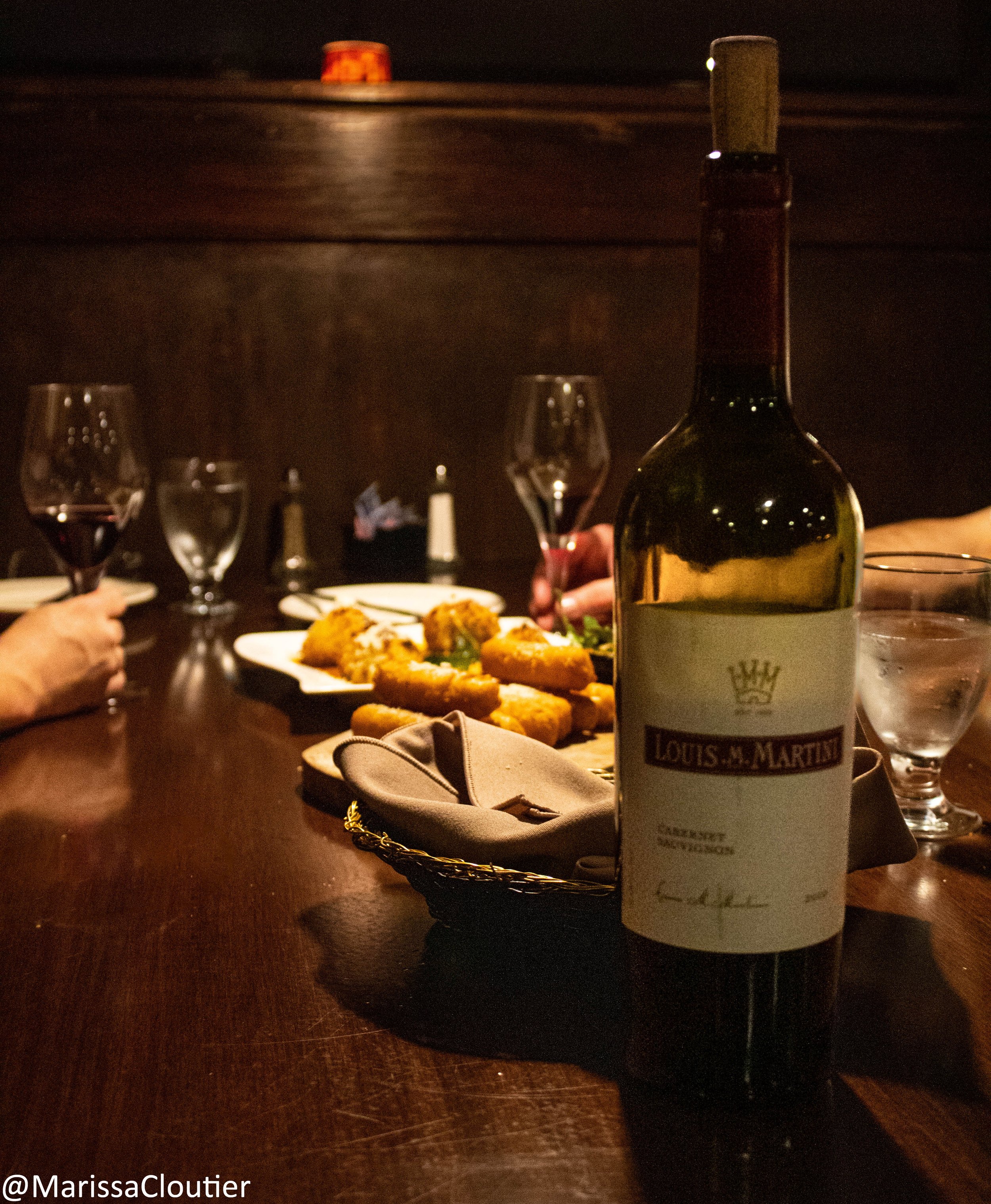 A dining table with a bottle of Louis M. Martini Cabernet Sauvignon, several glasses of red wine, plates of food, and people holding wine glasses in a dimly lit setting.