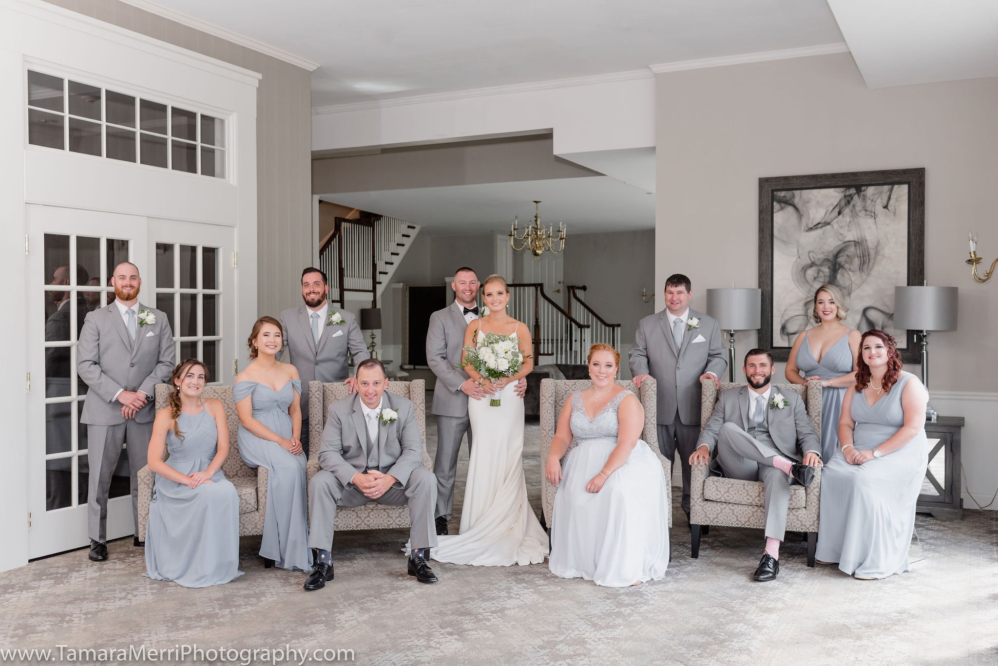 A wedding party of eleven people, including the bride in a white dress holding a bouquet and ten guests in matching gray suits and light blue dresses, is posed in a spacious living room with a staircase, large artwork, lamps, and glass doors.