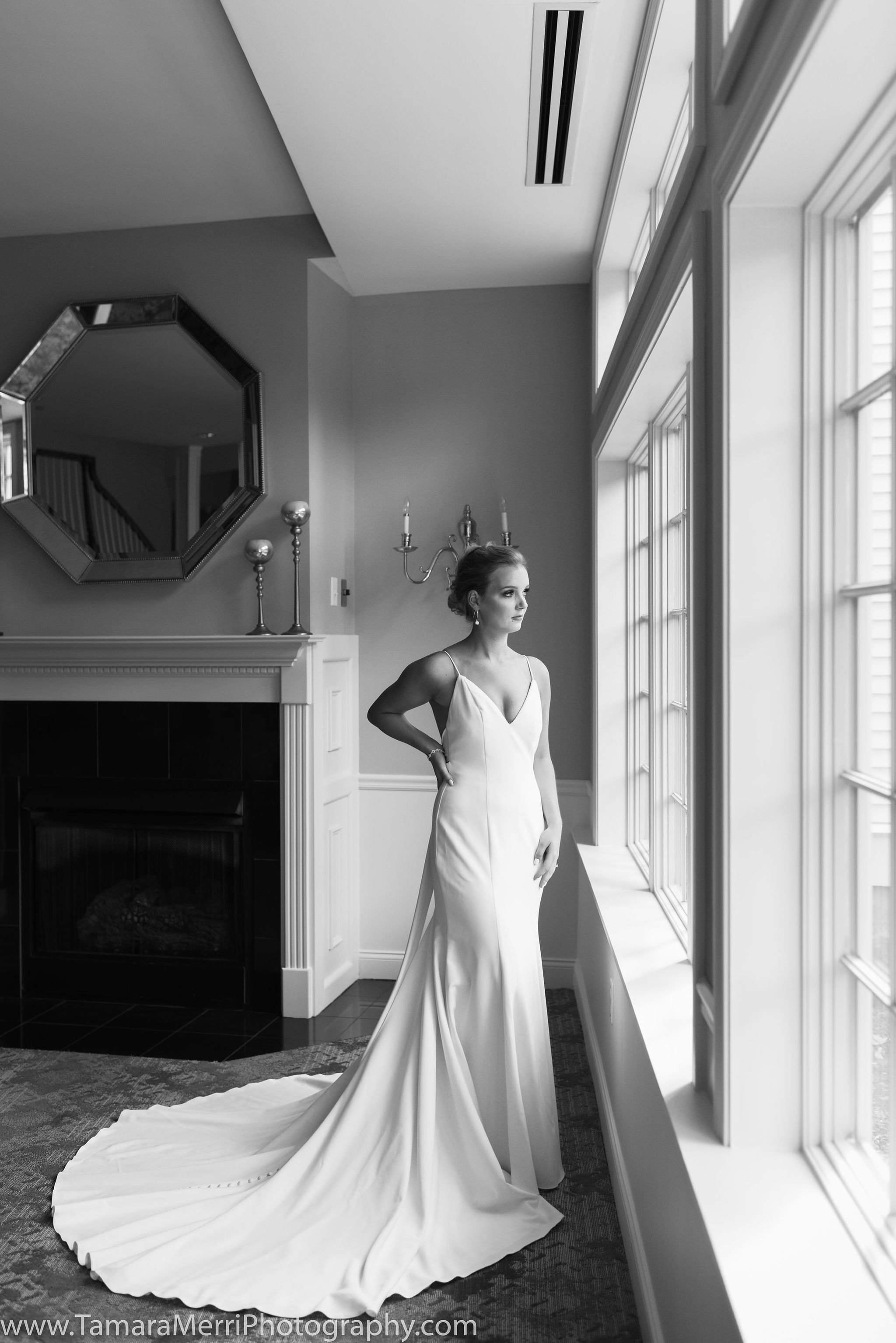 A woman in a wedding dress looking out a large window in a living room with decorative mirror and candelabra.
