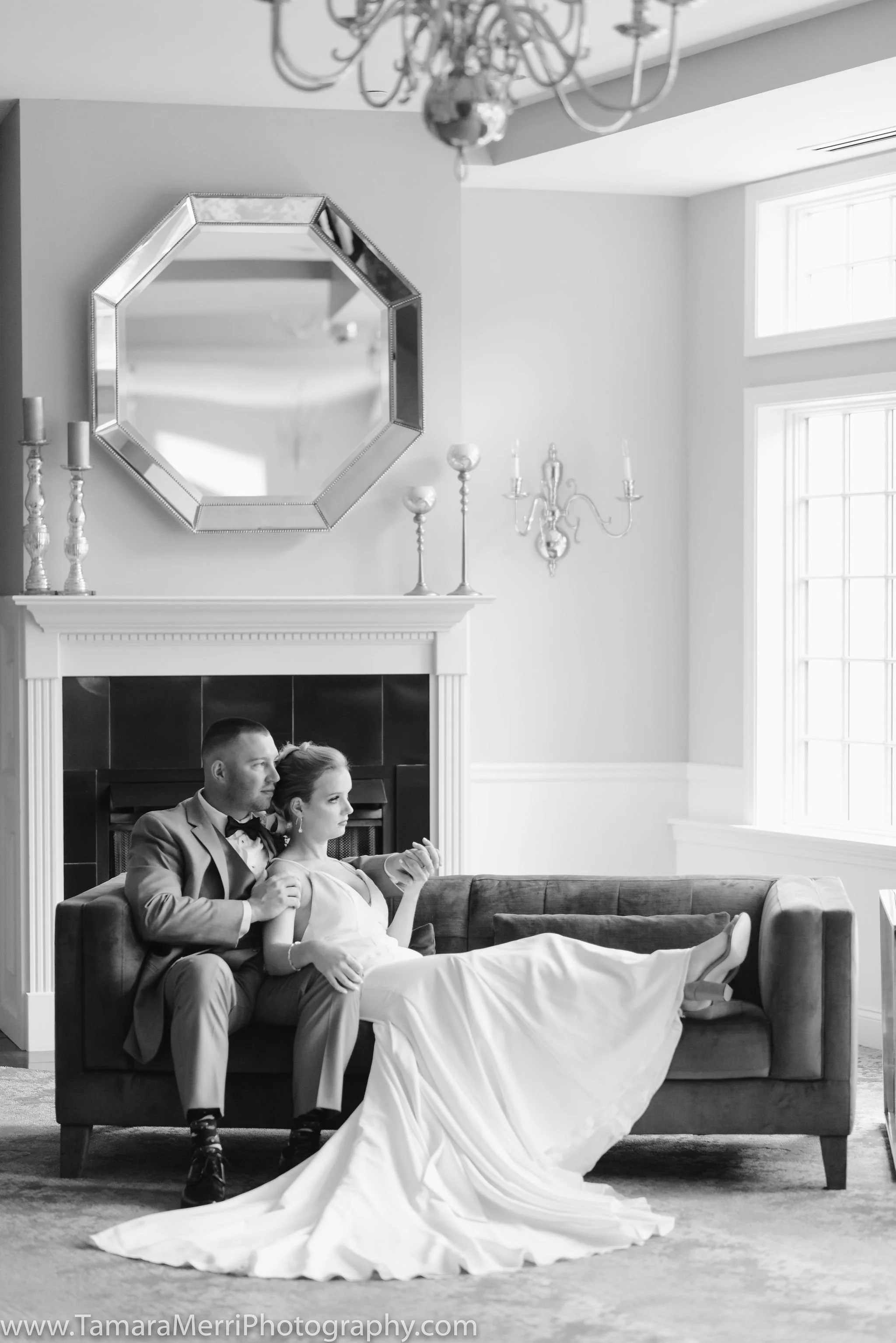 A black and white photo of a couple in wedding attire sitting on a sofa in a well-lit room with large windows and a decorative fireplace. The bride is reclining with her legs on the sofa, holding a smartphone, while the groom sits close behind her, h