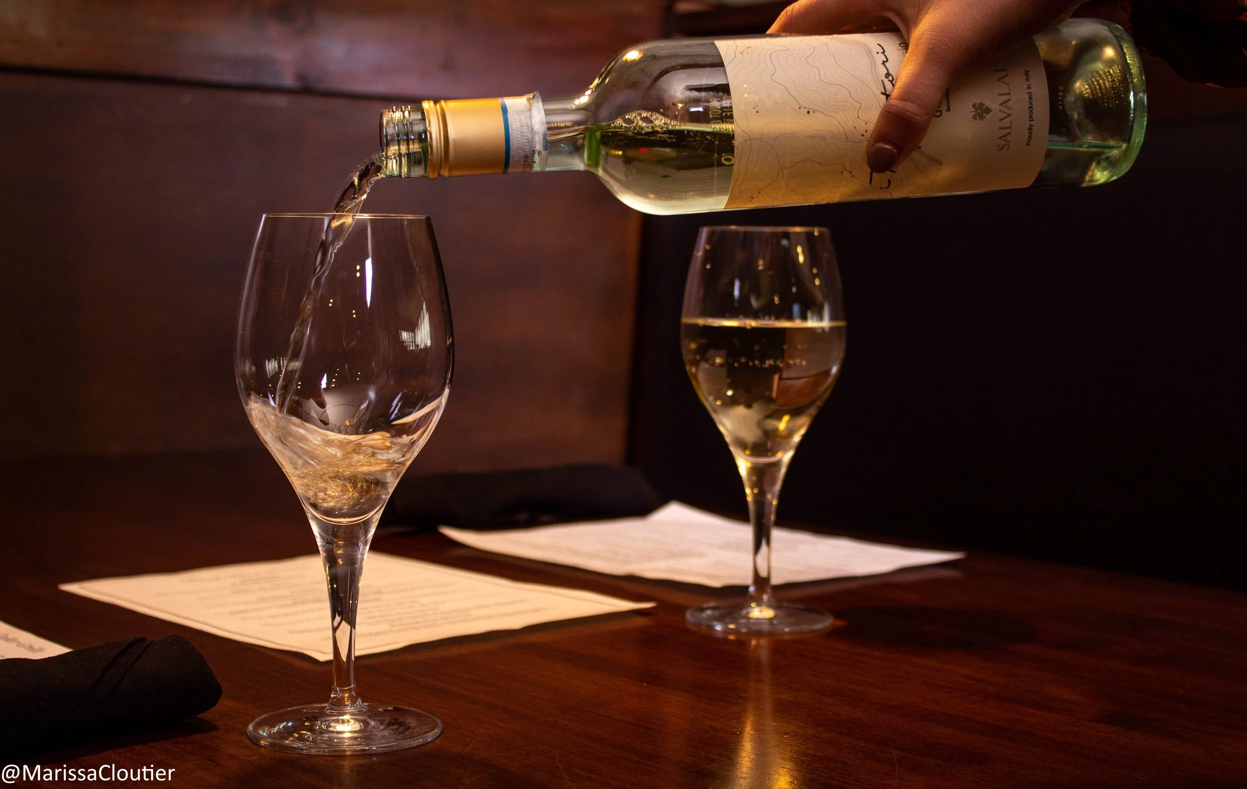 A person pouring white wine from a bottle into a wine glass on a wooden table, with another filled wine glass and menus in the background.