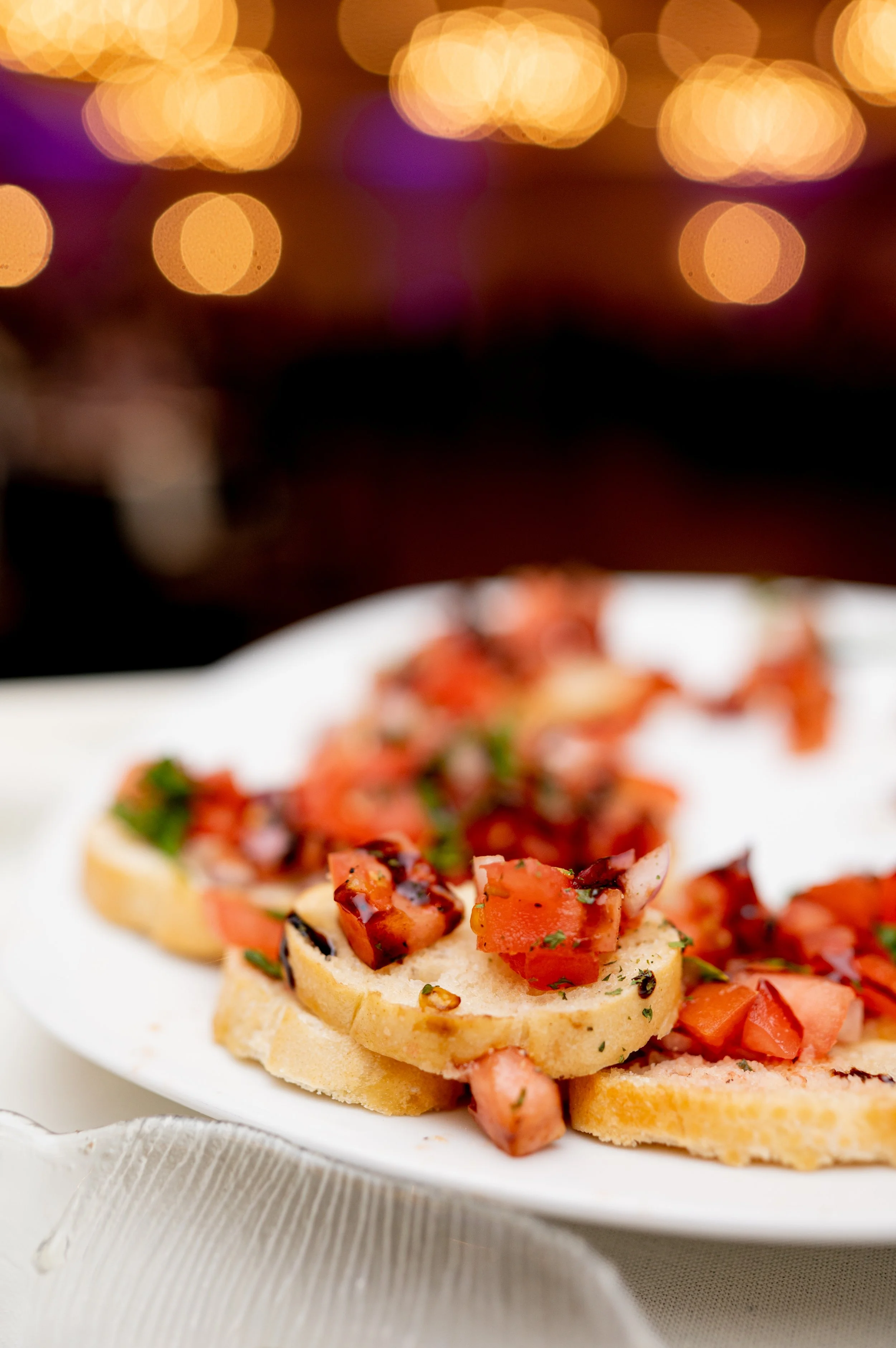 Close-up of tomato bruschetta on a white plate with blurred bokeh lights in the background.