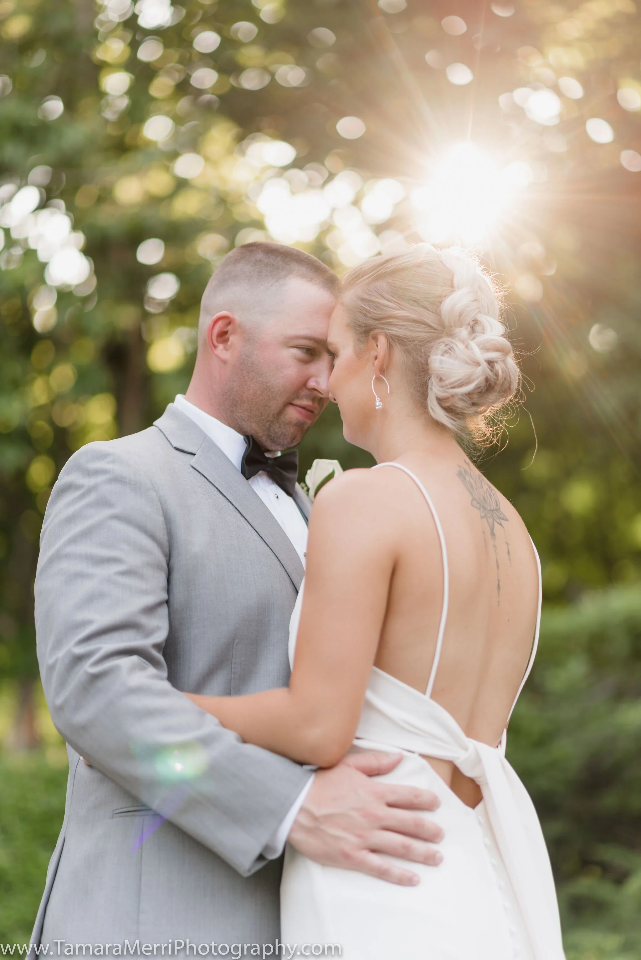 A bride and groom embrace outdoors, their foreheads touching, with the sun shining behind them and soft bokeh lights filtering through the trees.