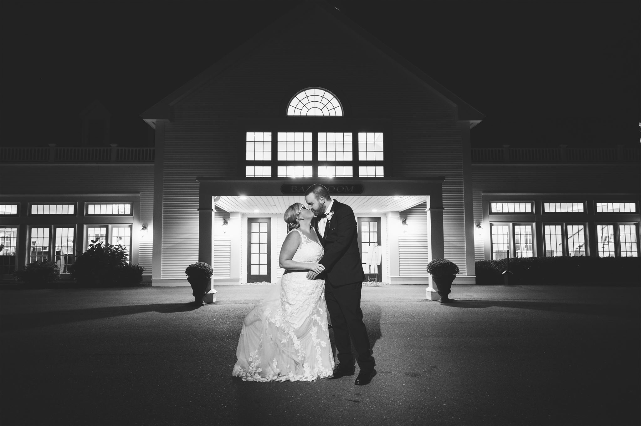 black and white photo of a bride and groom kissing outside a large lit building at night, with the bride in a wedding gown and the groom in a suit