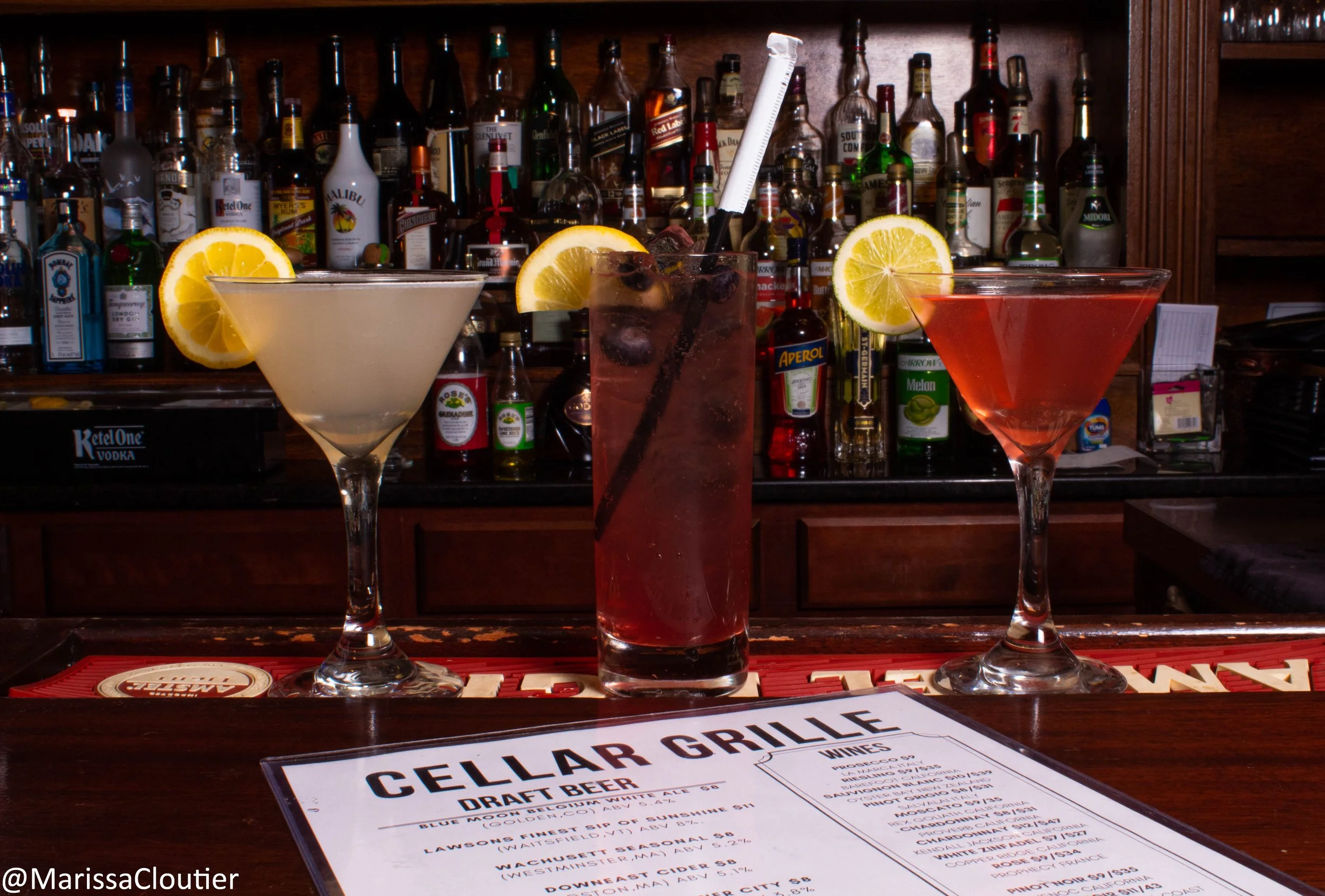 Three cocktails with lemon slices on a bar counter, with a bar back filled with various bottles of liquor in the background.