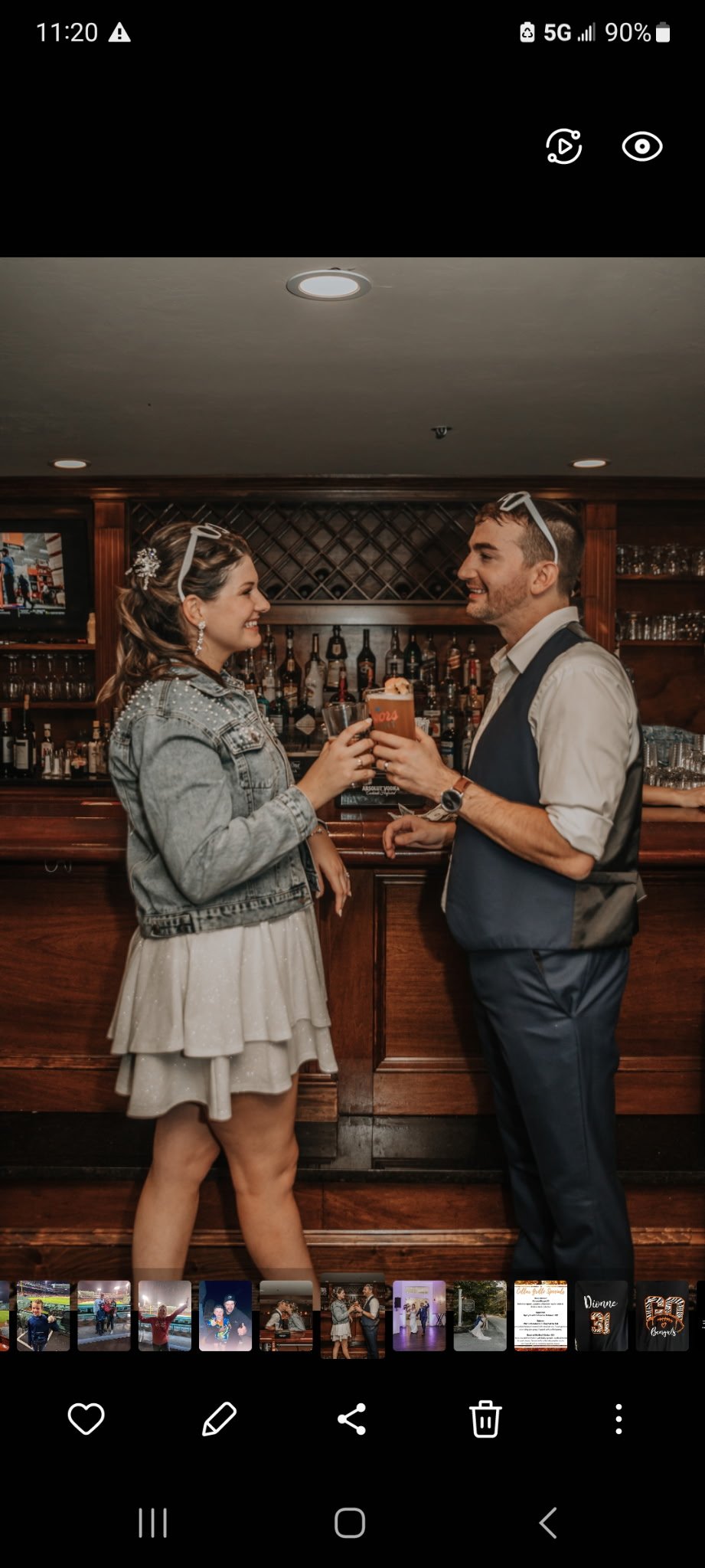 A man and woman are in a bar, holding drinks and looking at each other, smiling. The woman has wavy hair, is wearing a denim jacket, a short dress, and some accessories. The man has short hair, is wearing a white shirt with a vest, and sunglasses on his head. There are bottles of alcohol on shelves behind them.