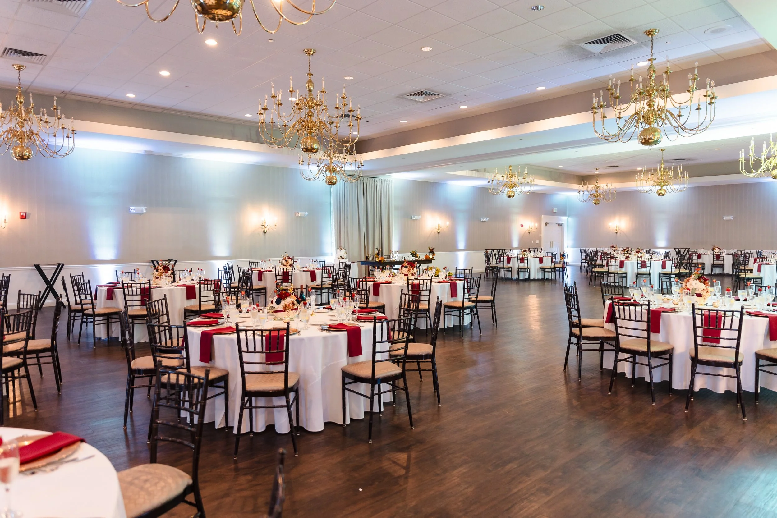 A banquet hall decorated for a wedding or event, with round tables covered in white tablecloths, red napkins, floral centerpieces, and black wooden chairs with beige cushions. The hall has wooden floors, beige walls, and multiple gold chandeliers hanging from the ceiling.