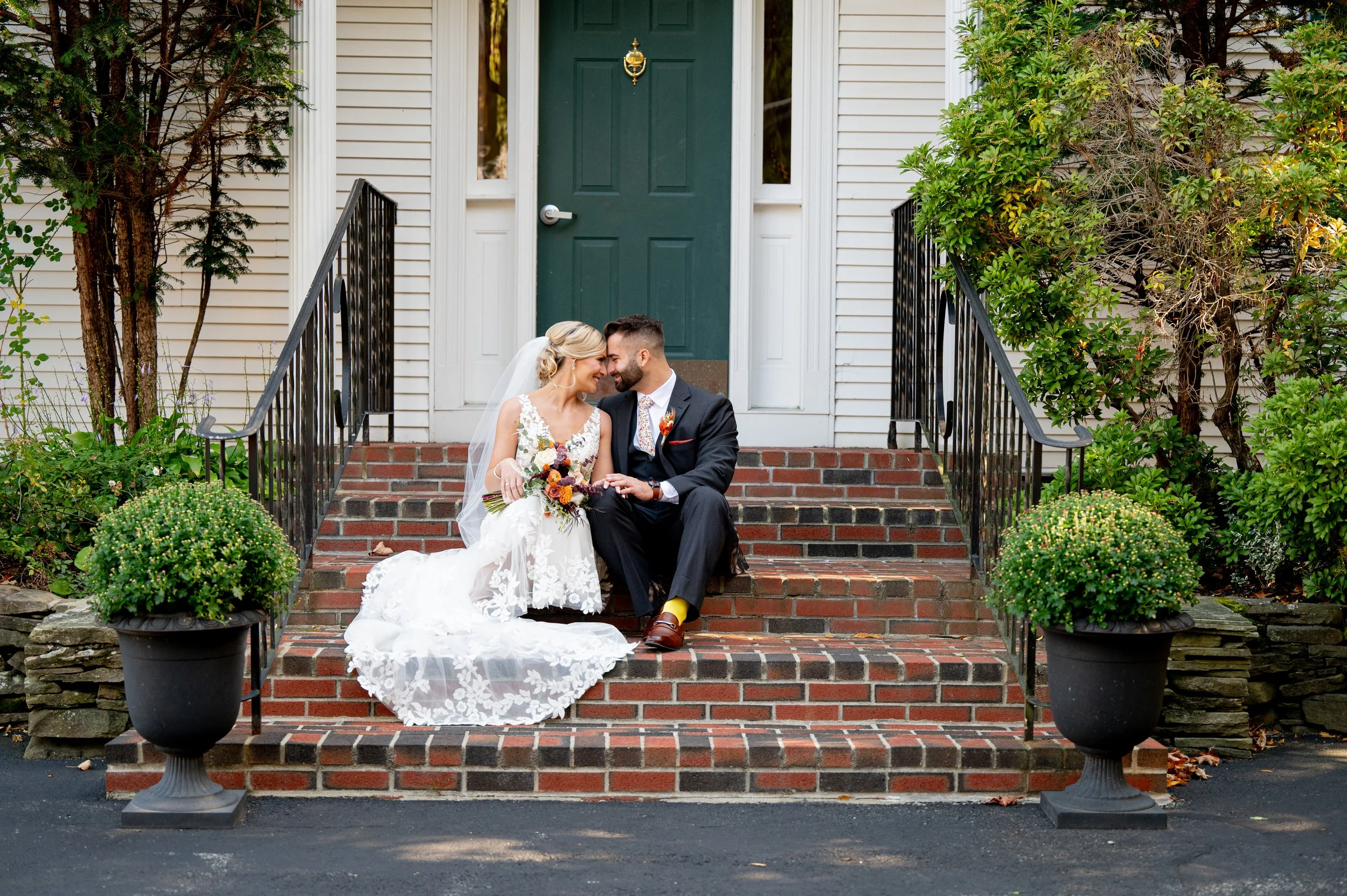 A bride and groom sitting on brick stairs in front of a house, smiling and touching foreheads, with the bride holding a colorful bouquet.